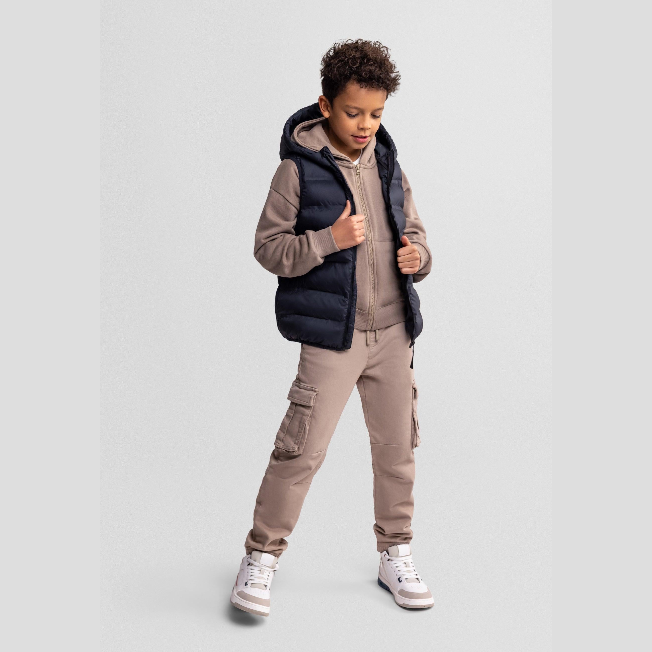 Boy in dynamic pose wearing black quilted waistcoat over beige tracksuit, hands pulling vest open, against light grey studio background