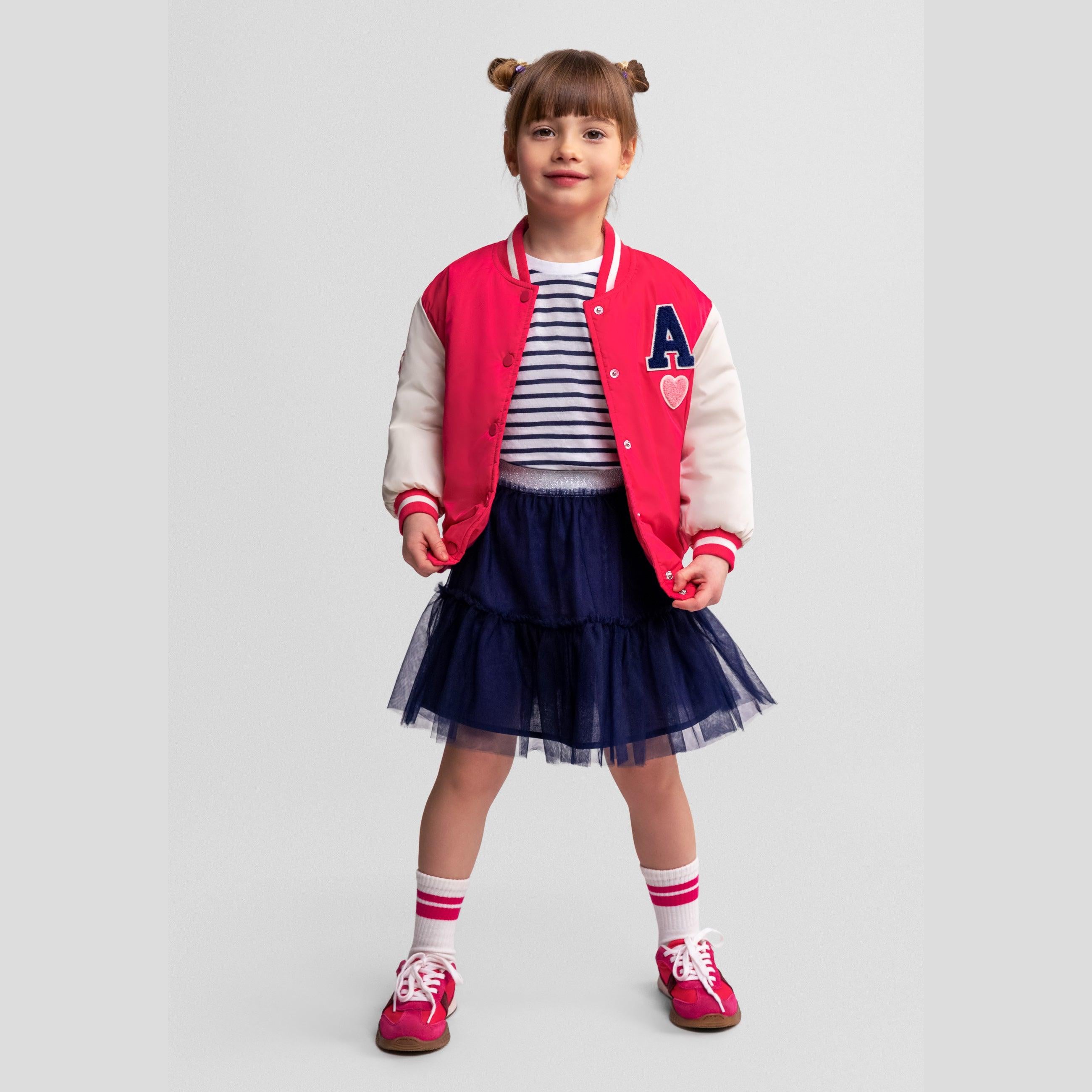 Front view of girl in pink varsity bomber jacket with letter A patch, navy tulle skirt and red sneakers on white background