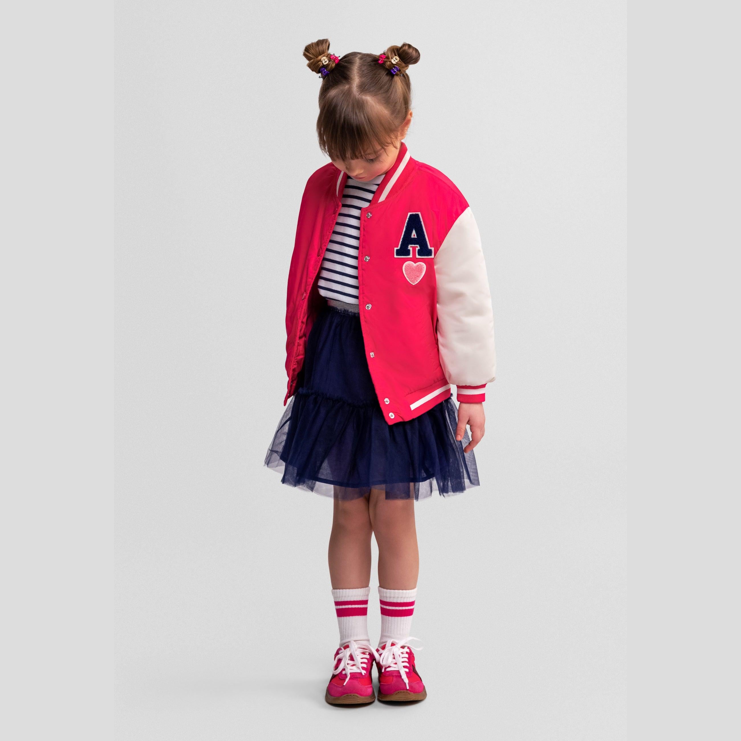 Front view of girl in pink varsity bomber jacket with letter A patch, navy tulle skirt and red sneakers on white background