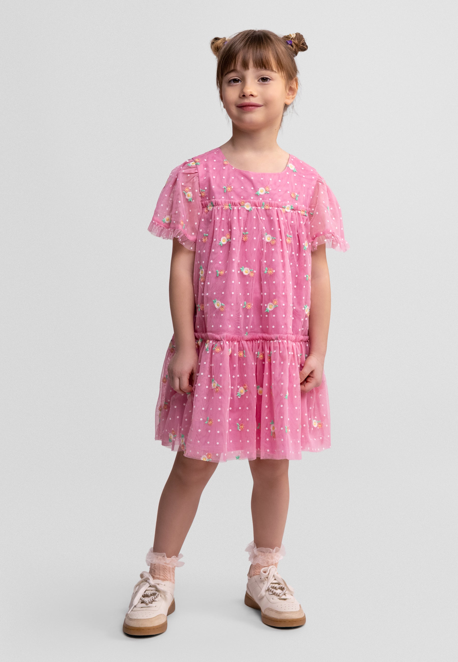 Front view of girl wearing pink tulle dress with floral embroidery, flutter sleeves and tiered skirt, styled with sneakers against white background