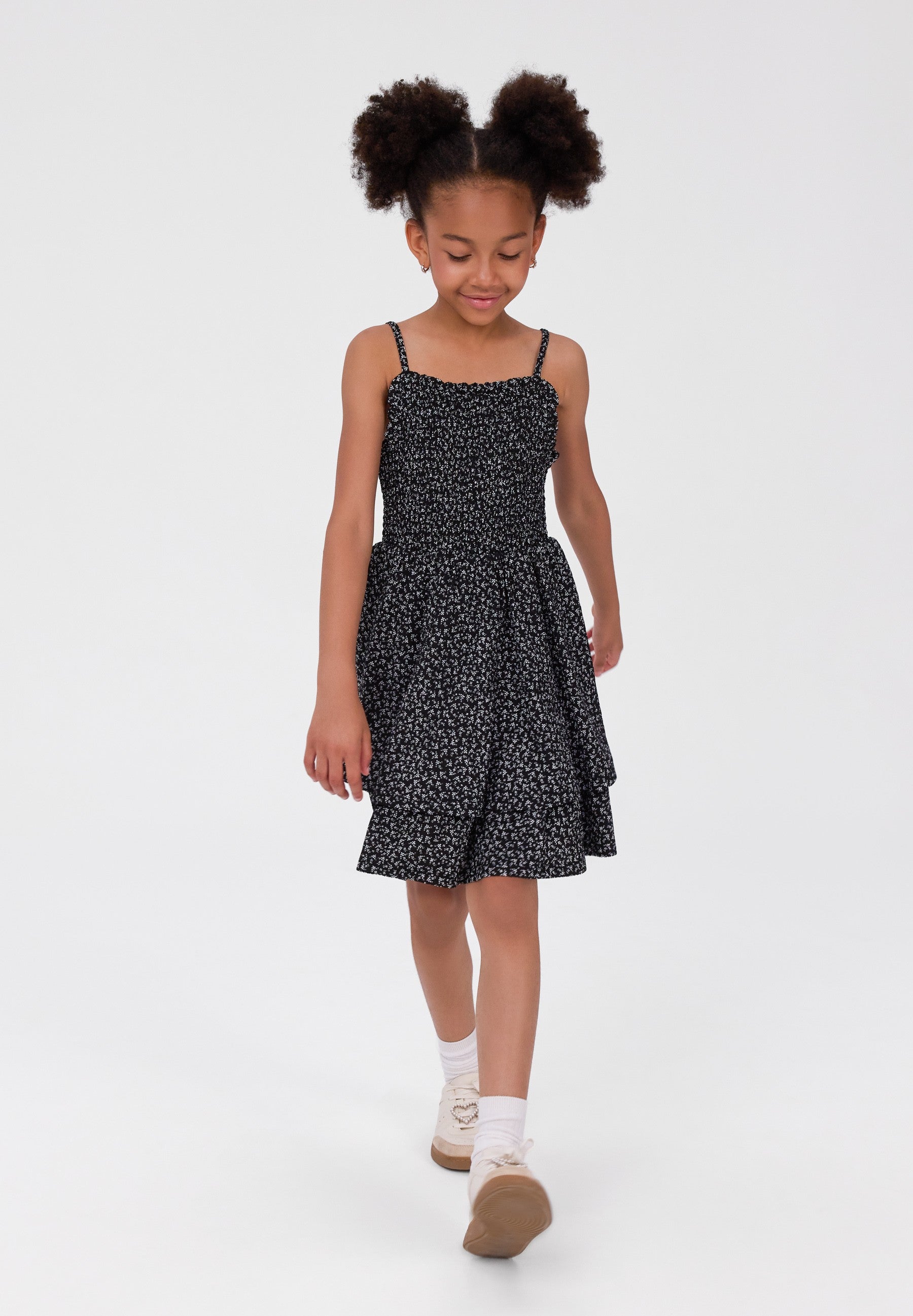A young girl with two high buns walks forward wearing a black spaghetti-strap dress with a small white floral print, white socks, and brown shoes against a plain white studio background.