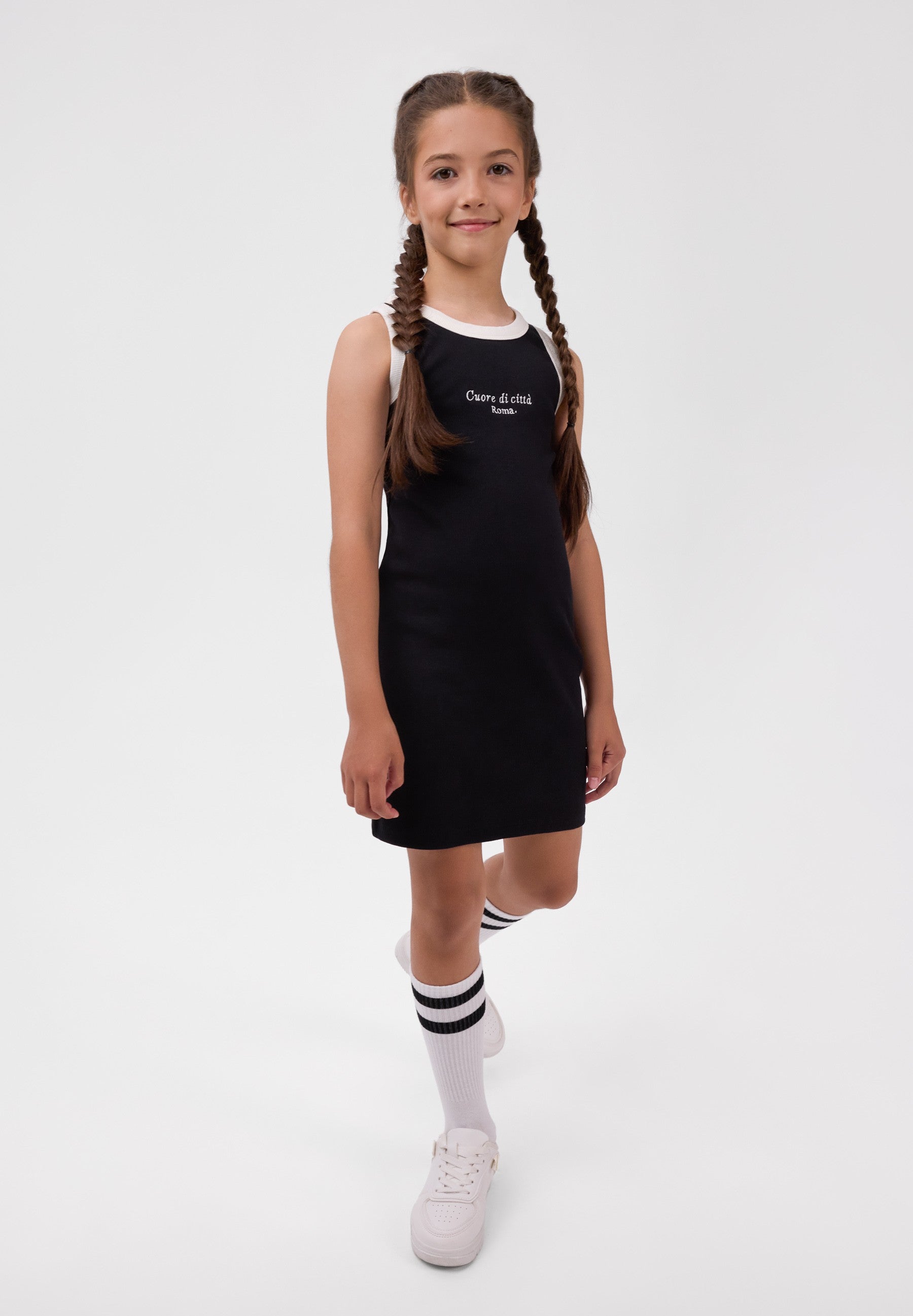 A girl wearing a black, form-fitting, sleeveless rib-knit dress with white “Cuore di città Roma” embroidery and white trim.