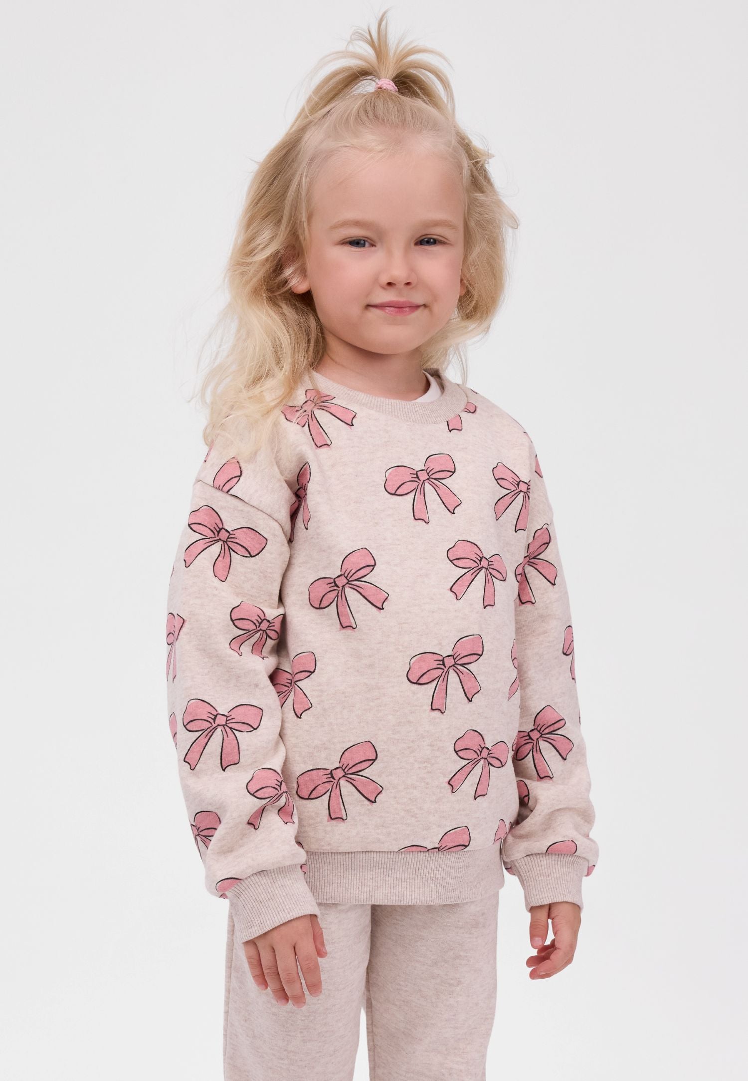 A young girl with blonde hair in a high ponytail wearing a light beige crewneck sweatshirt with an all-over pink bow pattern, paired with matching beige pants, against a plain light gray background.