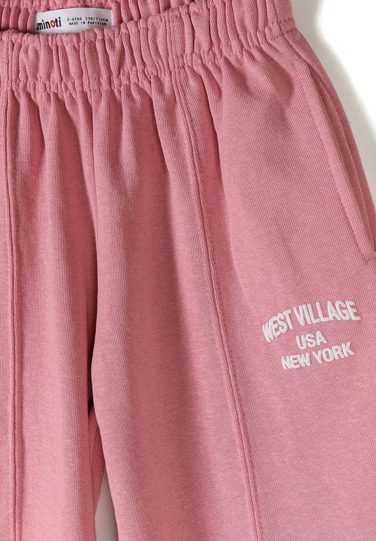 A close-up of dusty pink sweatpants featuring an elastic waistband and white embroidered text on the leg that reads 'WEST VILLAGE USA NEW YORK'.