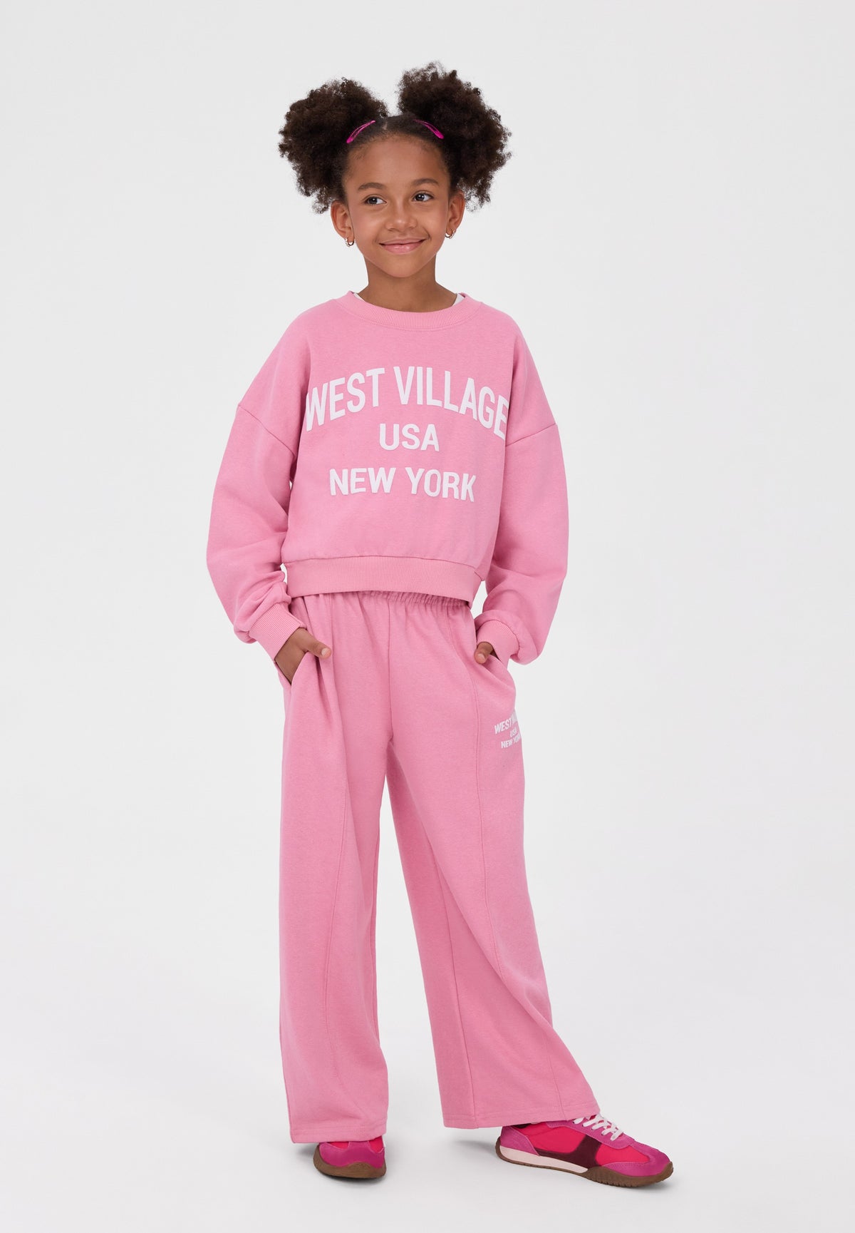 A young girl with her hair in two buns stands against a light gray studio background wearing a matching pink sweatshirt and sweatpants set. The sweatshirt has 'WEST VILLAGE USA NEW YORK' printed in white across the chest, and she is wearing pink sneakers.