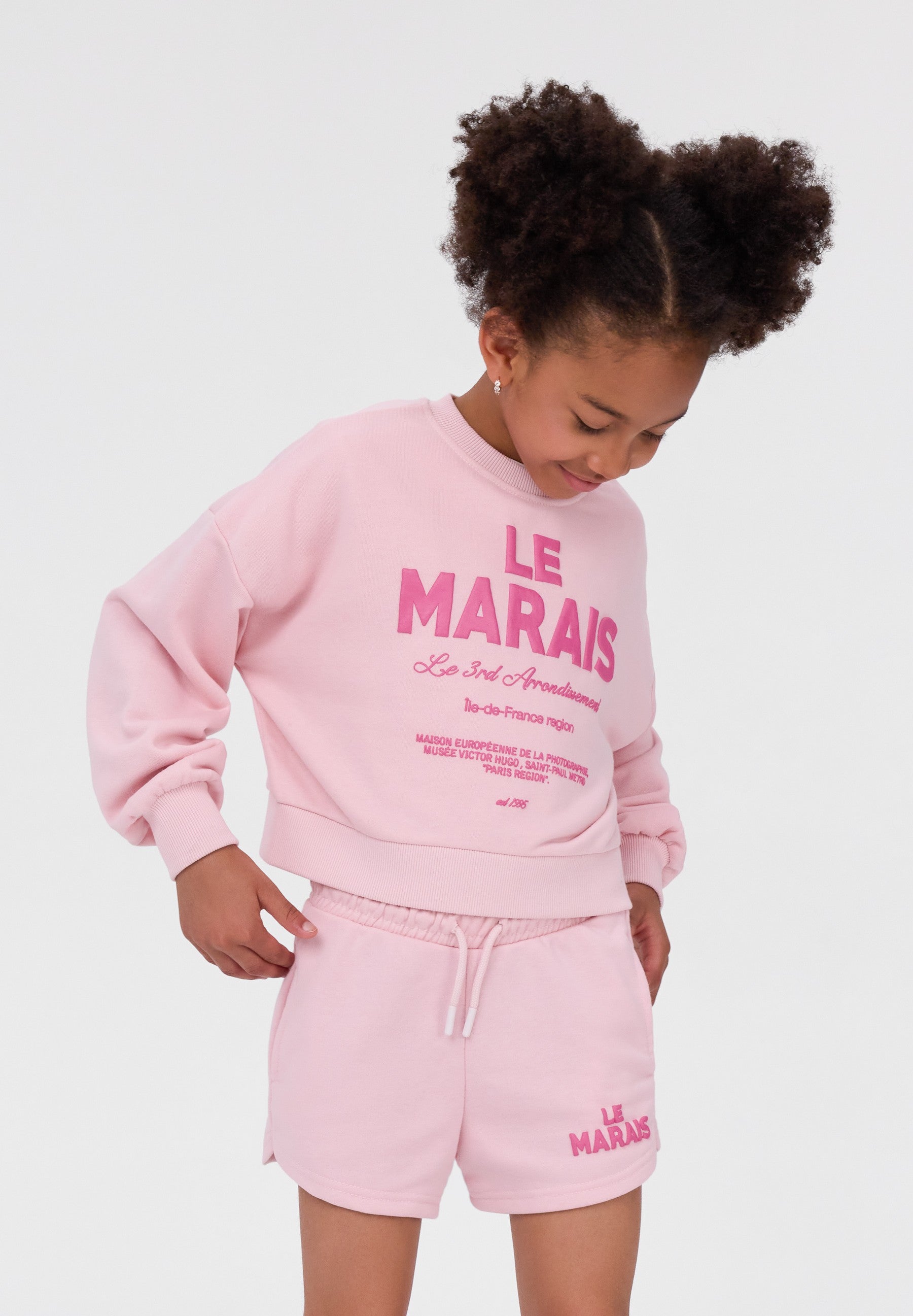 A young girl with curly hair styled in two buns is wearing a light pink sweatshirt and matching shorts set with 'LE MARAIS' printed in darker pink on the front and shorts.