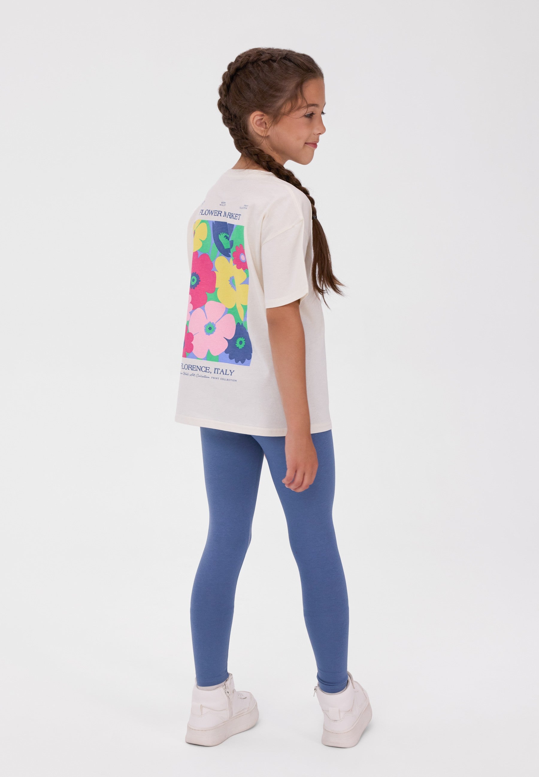 A young girl with a braided ponytail is shown from the back wearing a white t-shirt with a colorful floral graphic and blue leggings, standing against a plain white studio background.