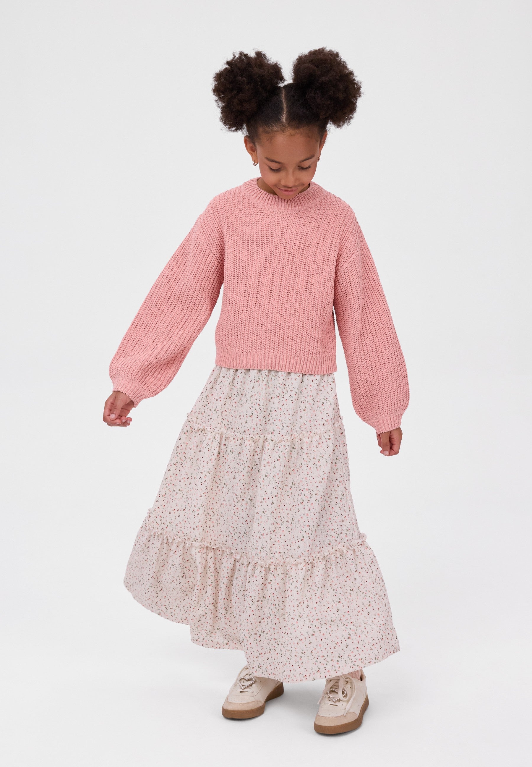 A young girl with two buns in her hair is wearing a pink ribbed knit mock-neck sweater and a light pink and white tiered floral skirt, standing against a plain white studio background.