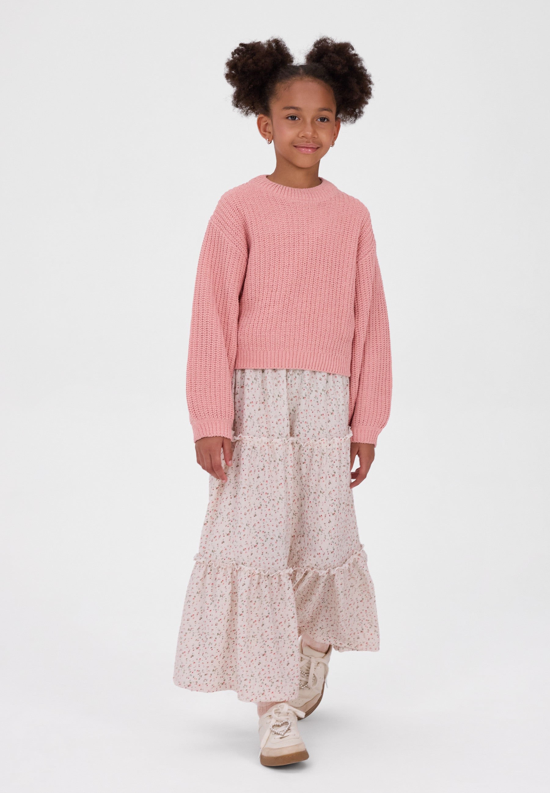 A young girl with two buns of hair stands against a light gray studio background wearing a pink ribbed knit sweater and a long tiered skirt with a small floral print.