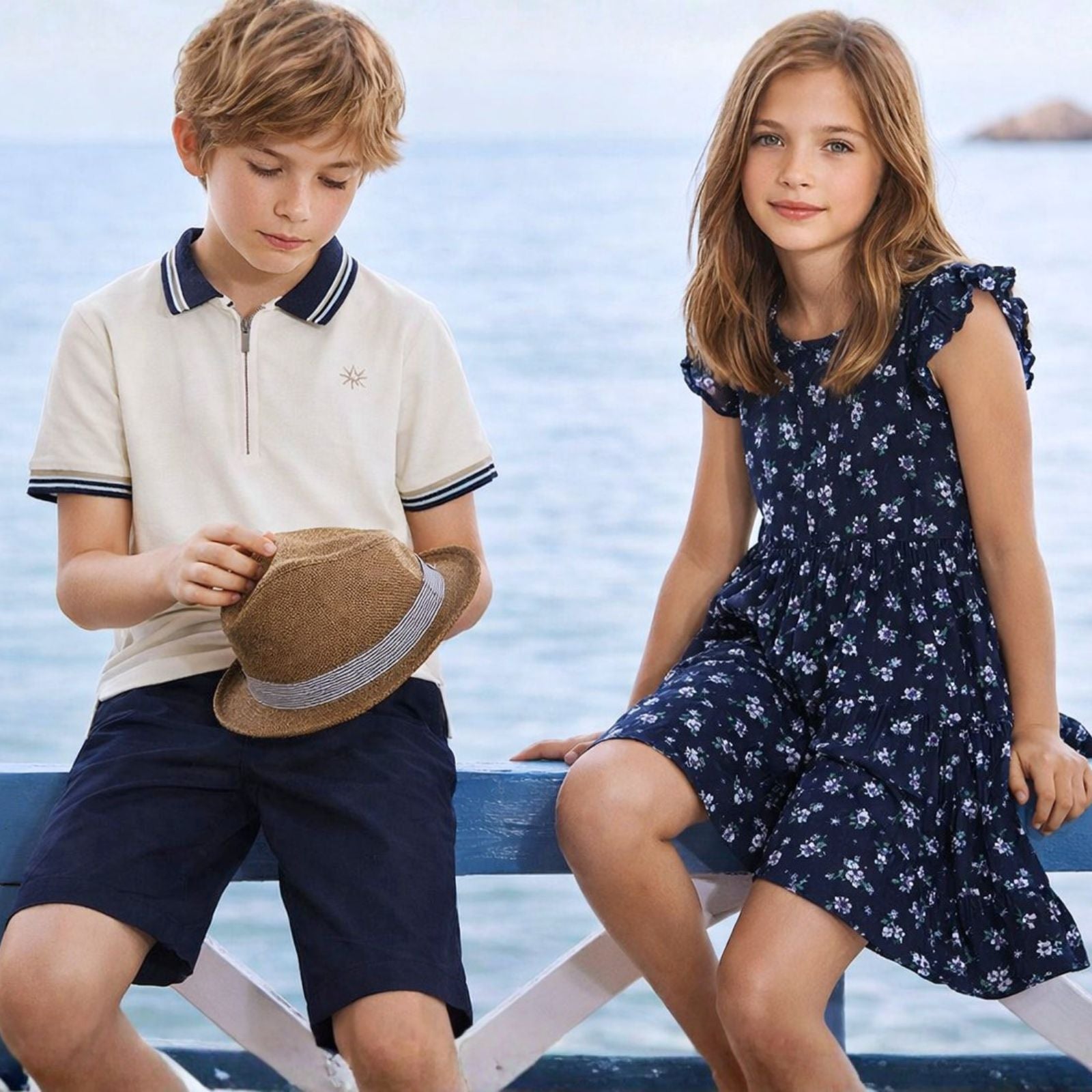 Two children, a boy and a girl, sitting by a body of water wearing summer clothing.