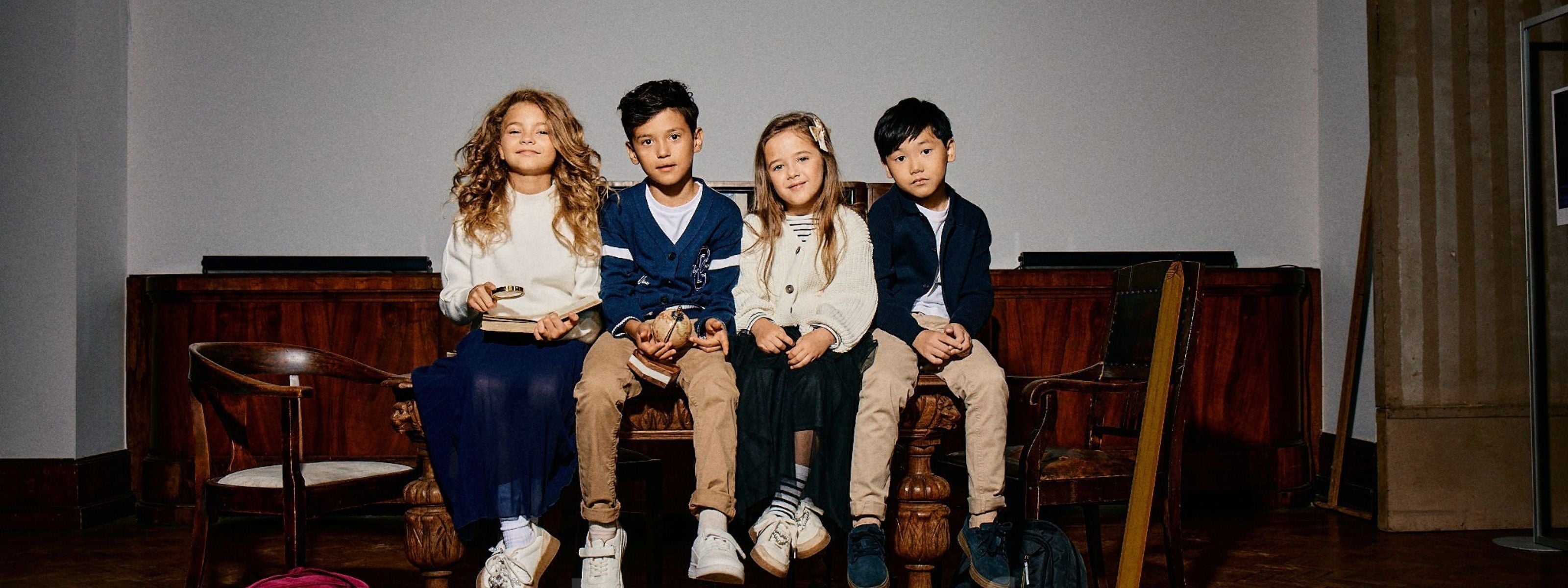 Four children sitting together on a wooden bench in an indoor setting, wearing coordinated outfits in neutral tones.