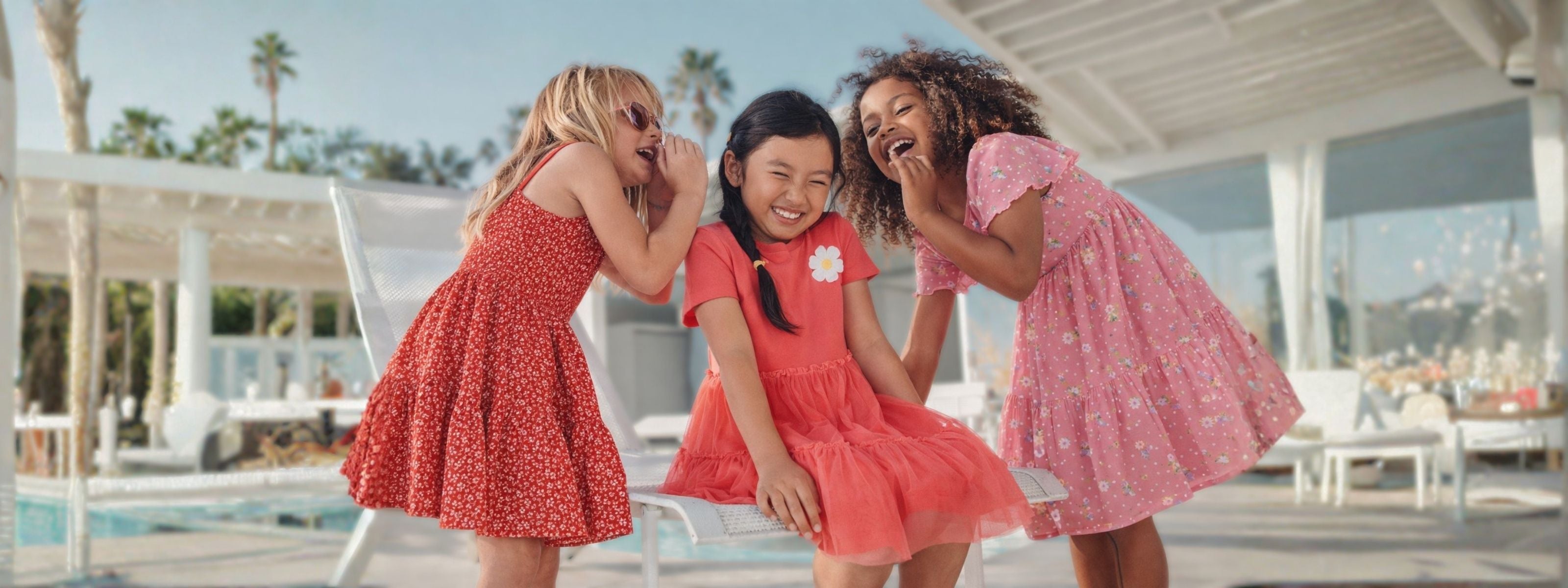 Three young girls in matching red dresses laughing by a poolside.