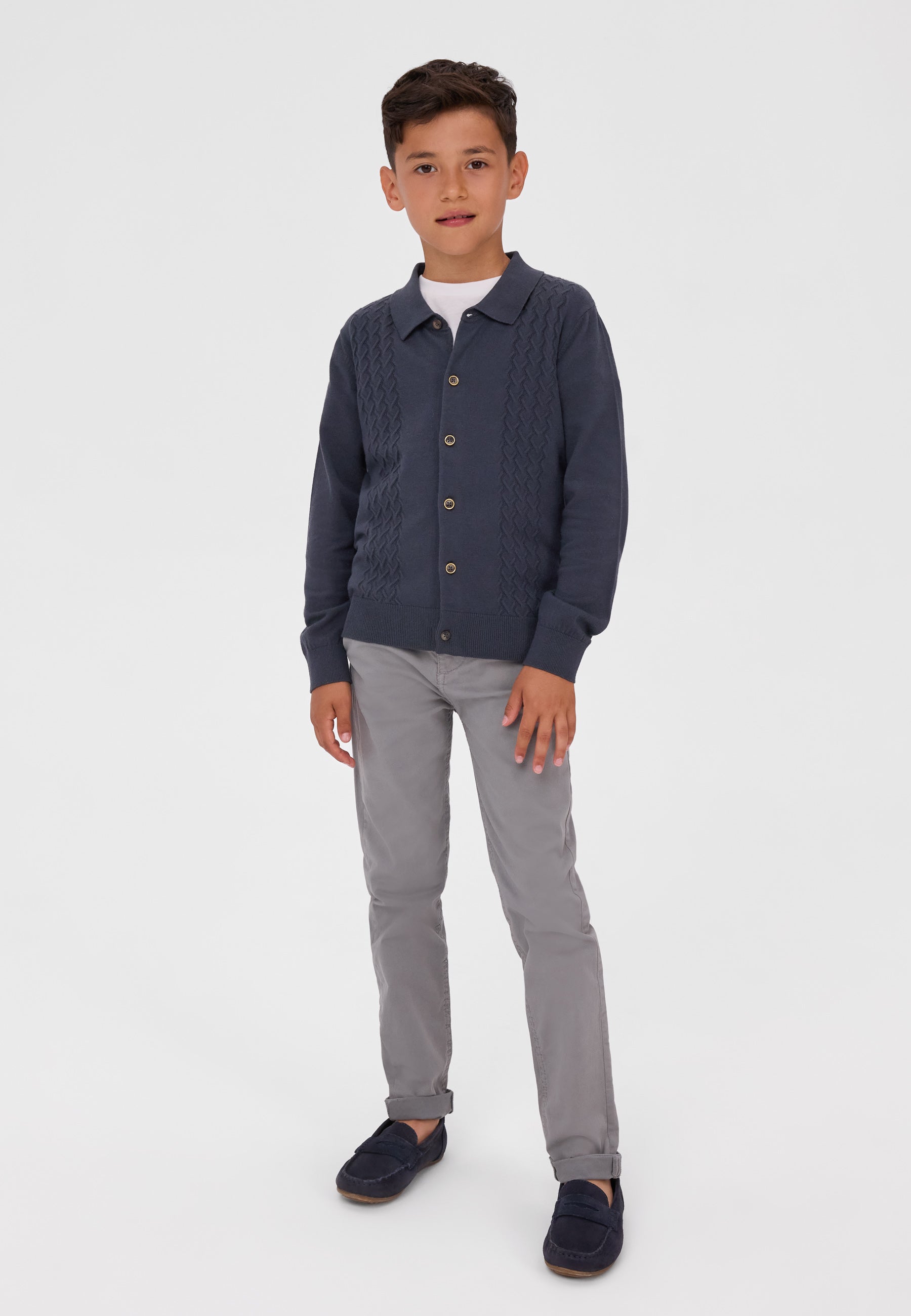 A young boy modeling a dark navy blue cable-knit button-up cardigan over a white t-shirt, paired with gray trousers and navy loafers, against a plain light gray studio background.