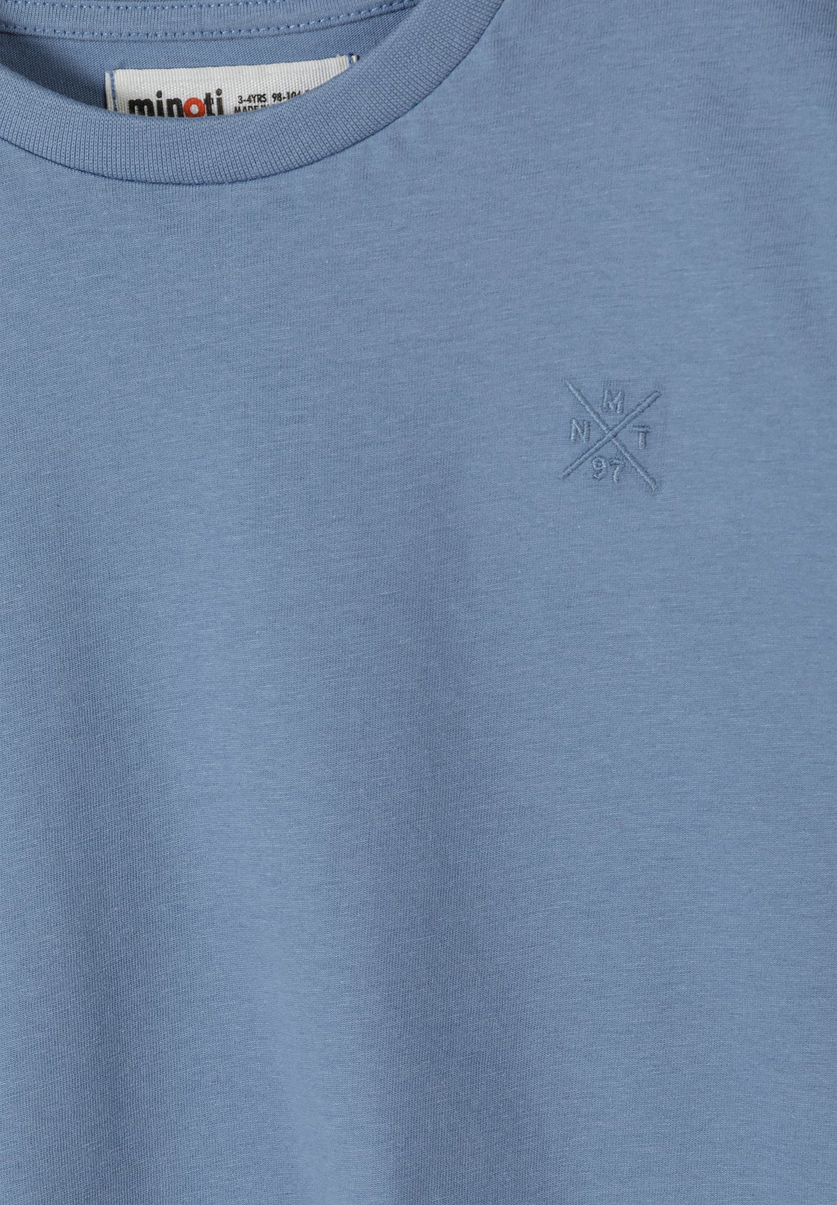 Close-up detail of blue cotton t-shirt showing subtle cross logo embossed on chest area with Minoti label visible at neckline