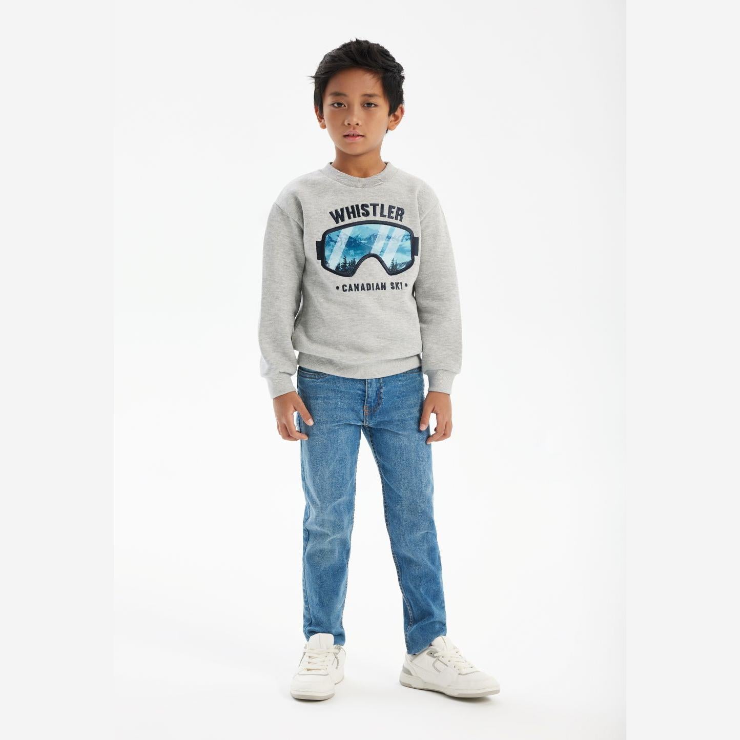 Young boy wearing grey crew neck sweatshirt with Whistler ski goggles graphic, paired with blue jeans and white sneakers, standing against white background