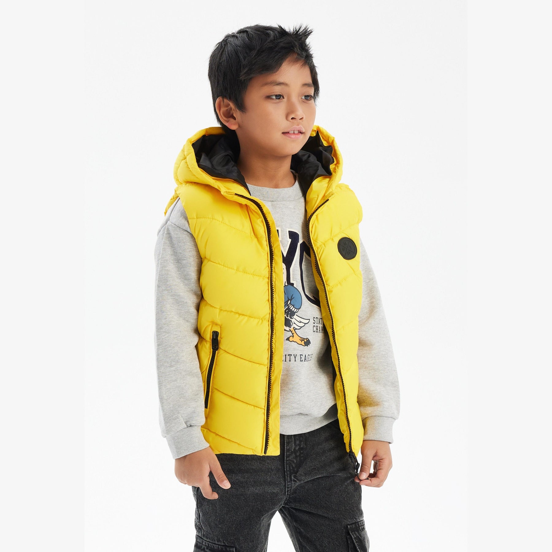 Boy wearing bright yellow padded gilet with black hood over grey sweatshirt and dark jeans against white background