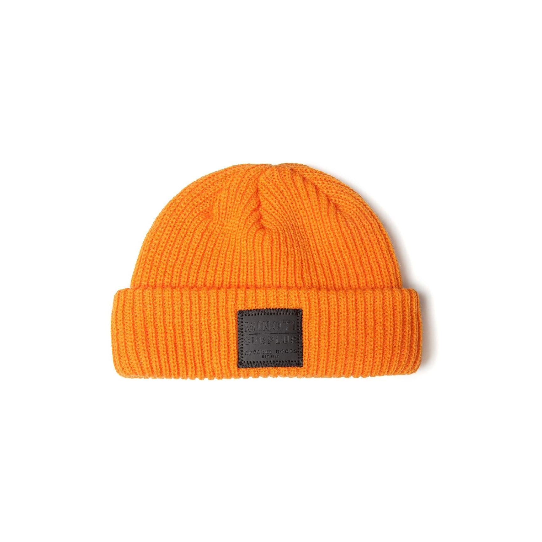 Close-up detail of dark grey rectangular leather patch on light grey ribbed beanie showing Minoti Surplus Apparel Goods Est. 1997 text with decorative stitching
