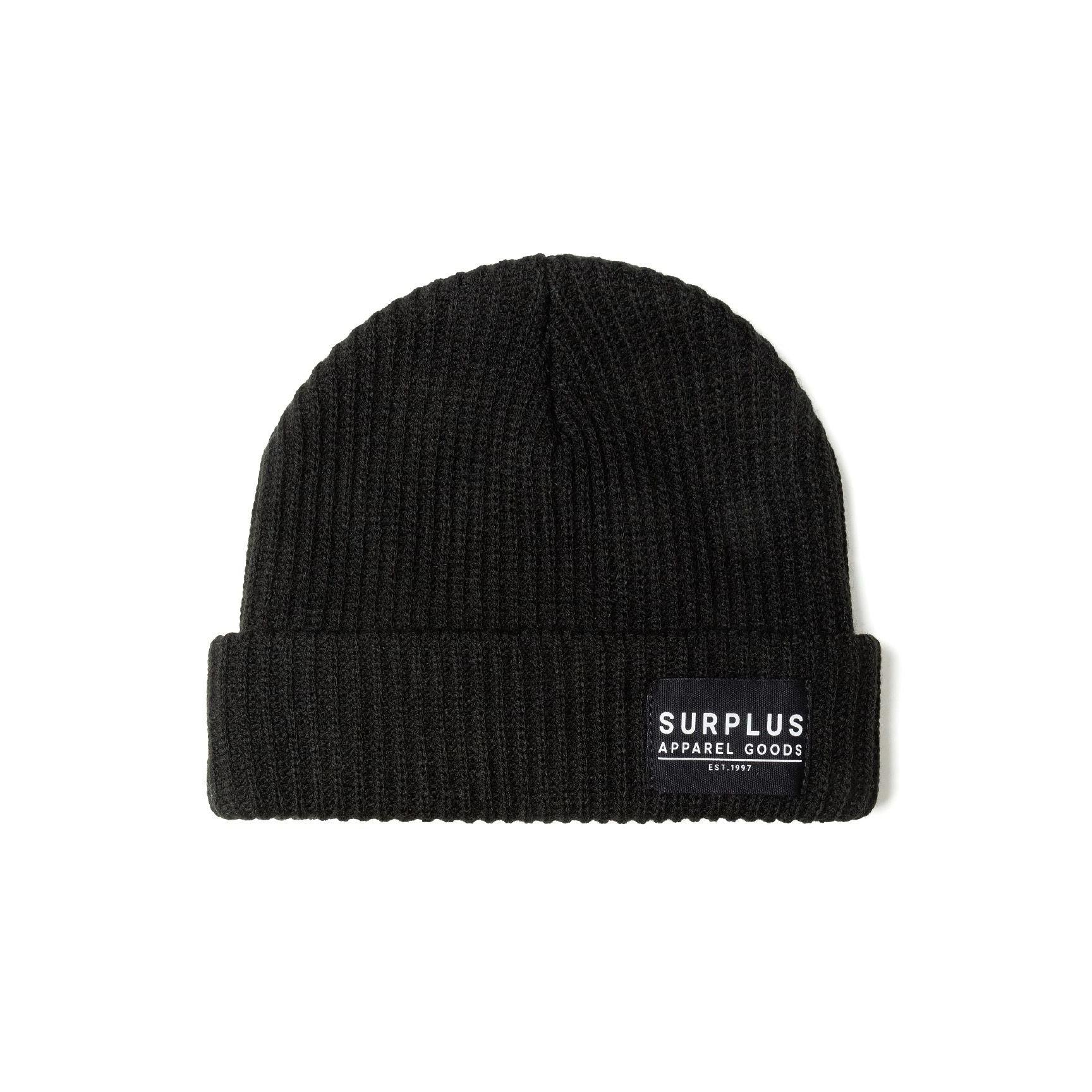 Close-up detail of black beanie showing SURPLUS APPAREL GOODS EST. 1997 woven label on ribbed knit fabric