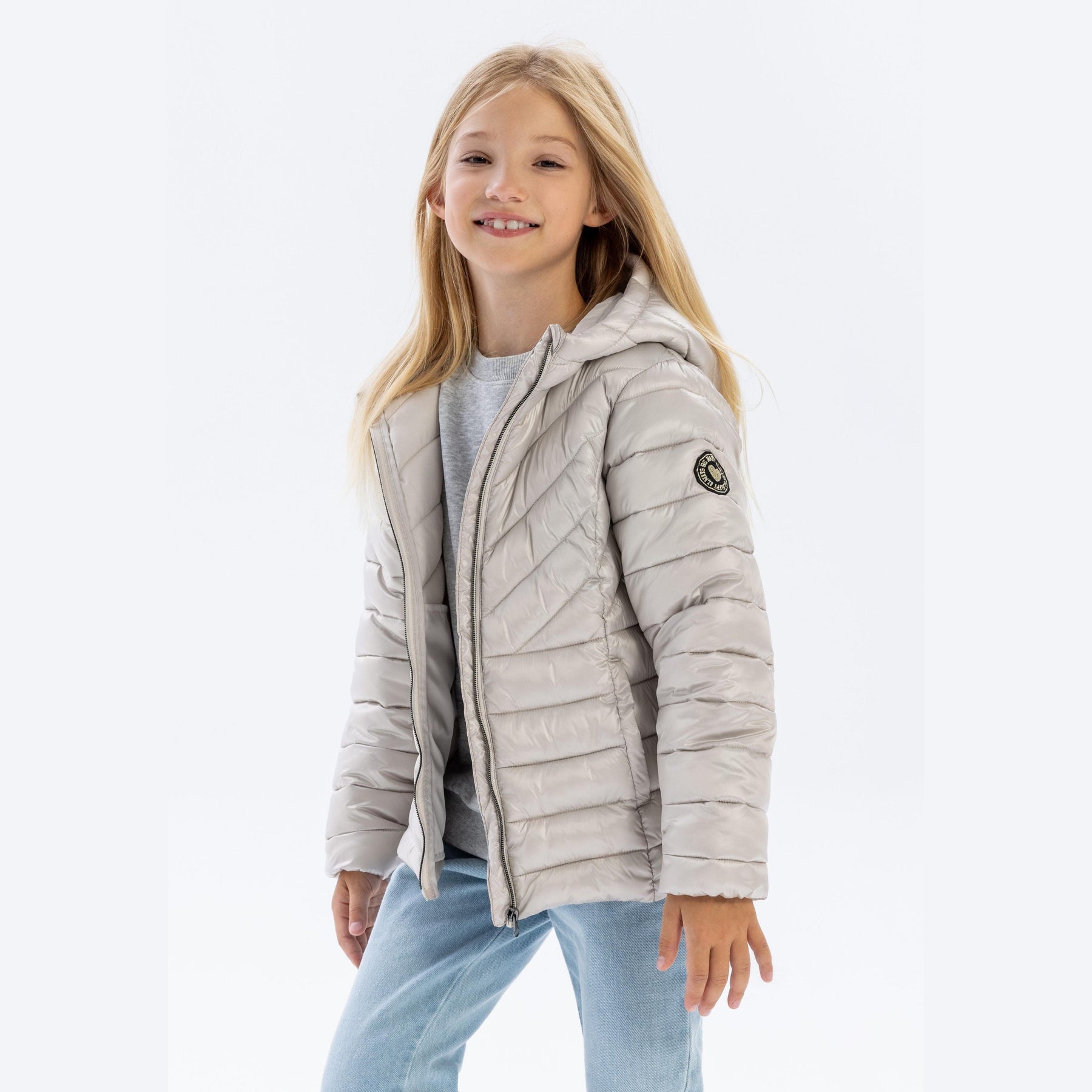 Young blonde girl wearing grey quilted jacket with hood up, smiling at camera, paired with light blue jeans against white background
