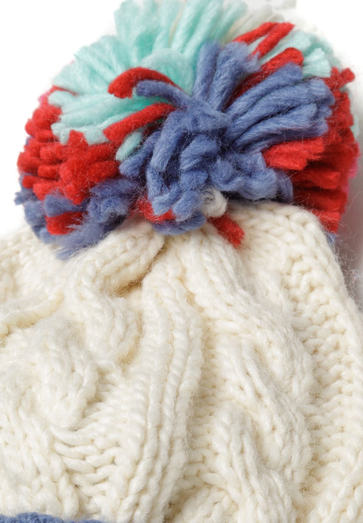Detail of a cream knitted hat with a colorful pom pom on top and a cable knit pattern.