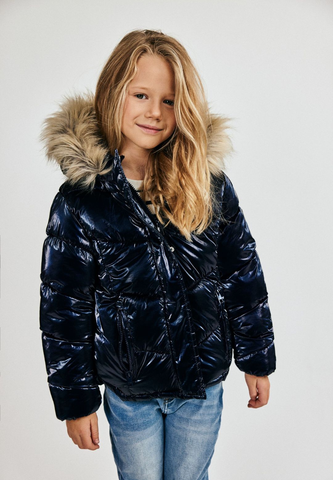 Girl wearing a glossy navy puffer jacket with beige fur-look hood trim and zipped side pockets.