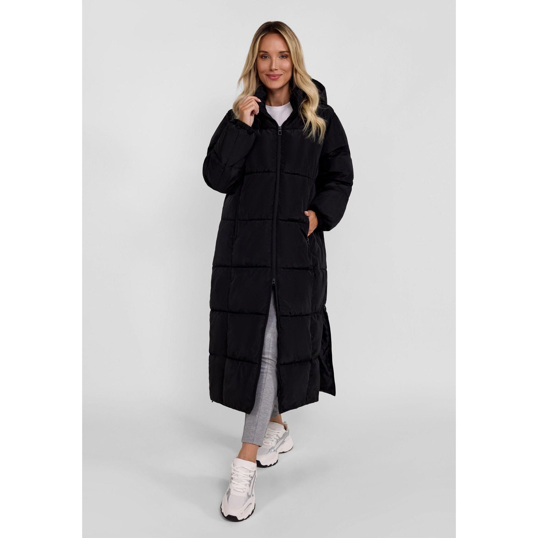 Blonde woman wearing long black quilted jacket with hood, paired with grey leggings and white sneakers, standing against white background