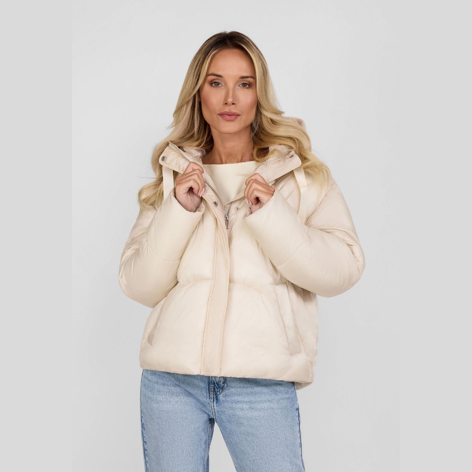 Woman wearing cream puffer jacket with hood up, paired with light blue jeans against white studio background