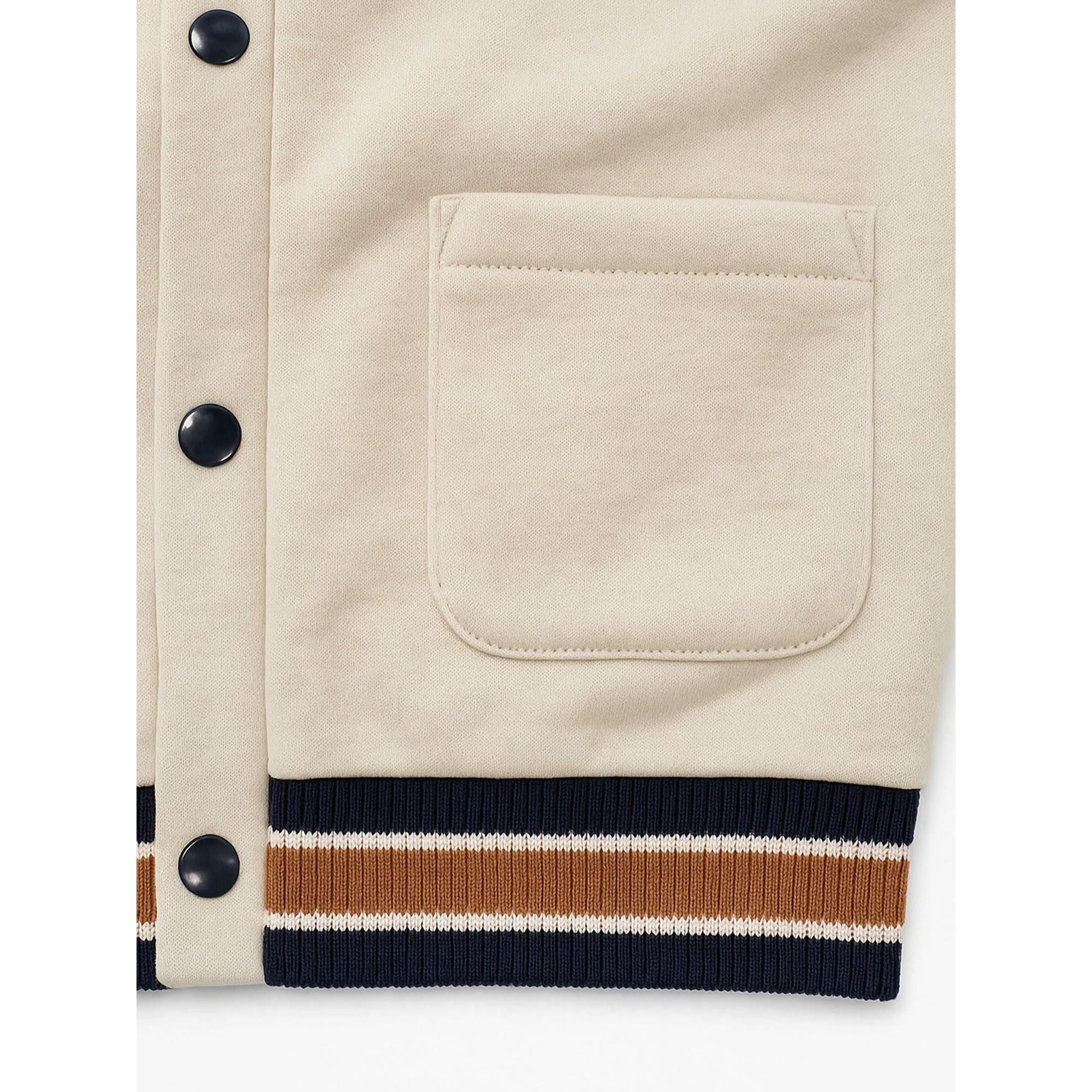 Close-up detail of cream cardigan showing chest pocket, navy contrast buttons and varsity-style striped ribbed hem trim
