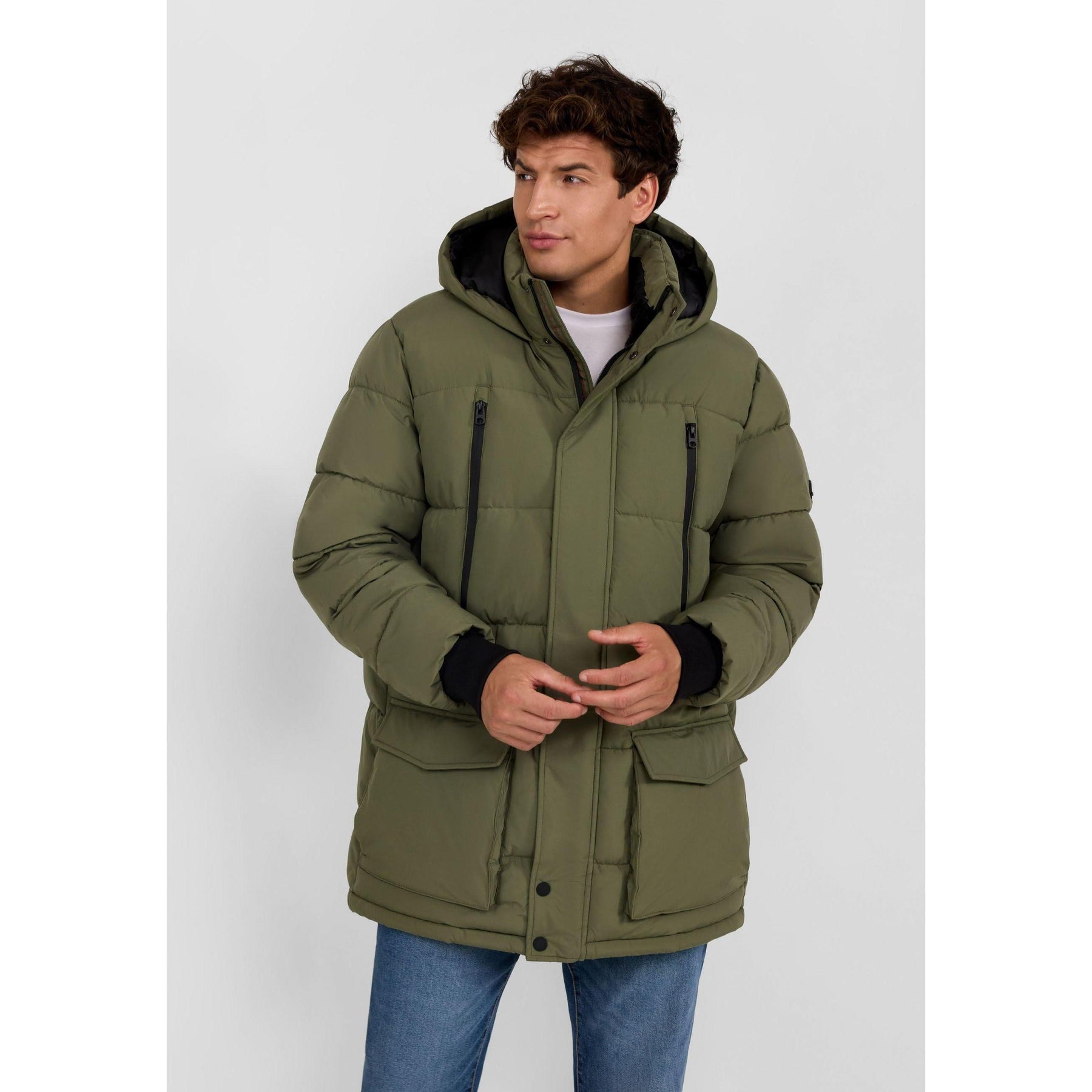 Young man wearing khaki quilted puffer jacket with hood and zip pockets, paired with blue jeans against light grey background