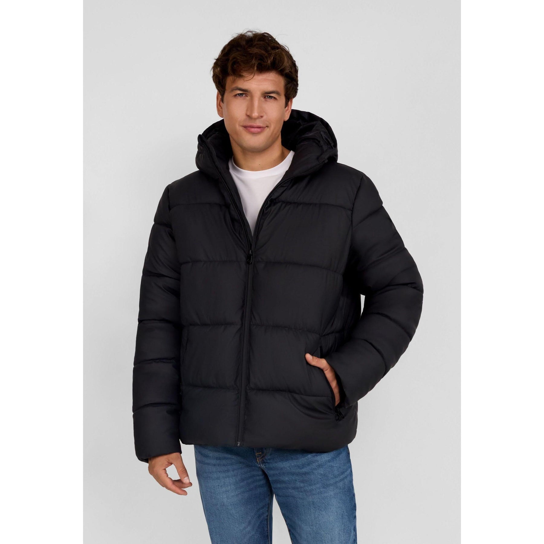 Young man wearing black quilted PU jacket with hood up, hands in pockets, standing against light grey background