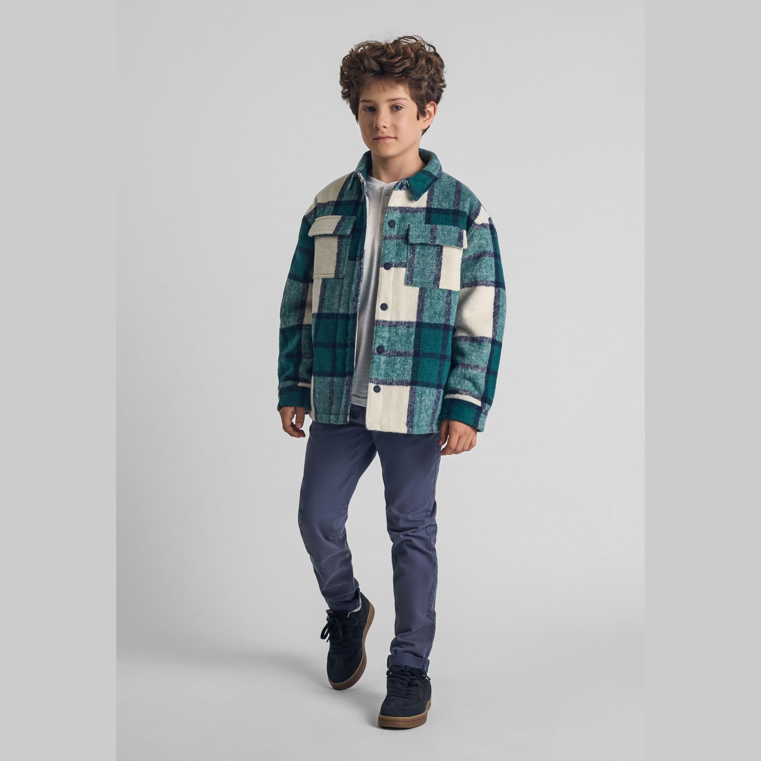 Young boy wearing green check padded shirt jacket with navy trousers and dark trainers, full body front view on light background