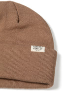 Detail of a ribbed beanie hat in brown with a fold-up cuff and logo badge.