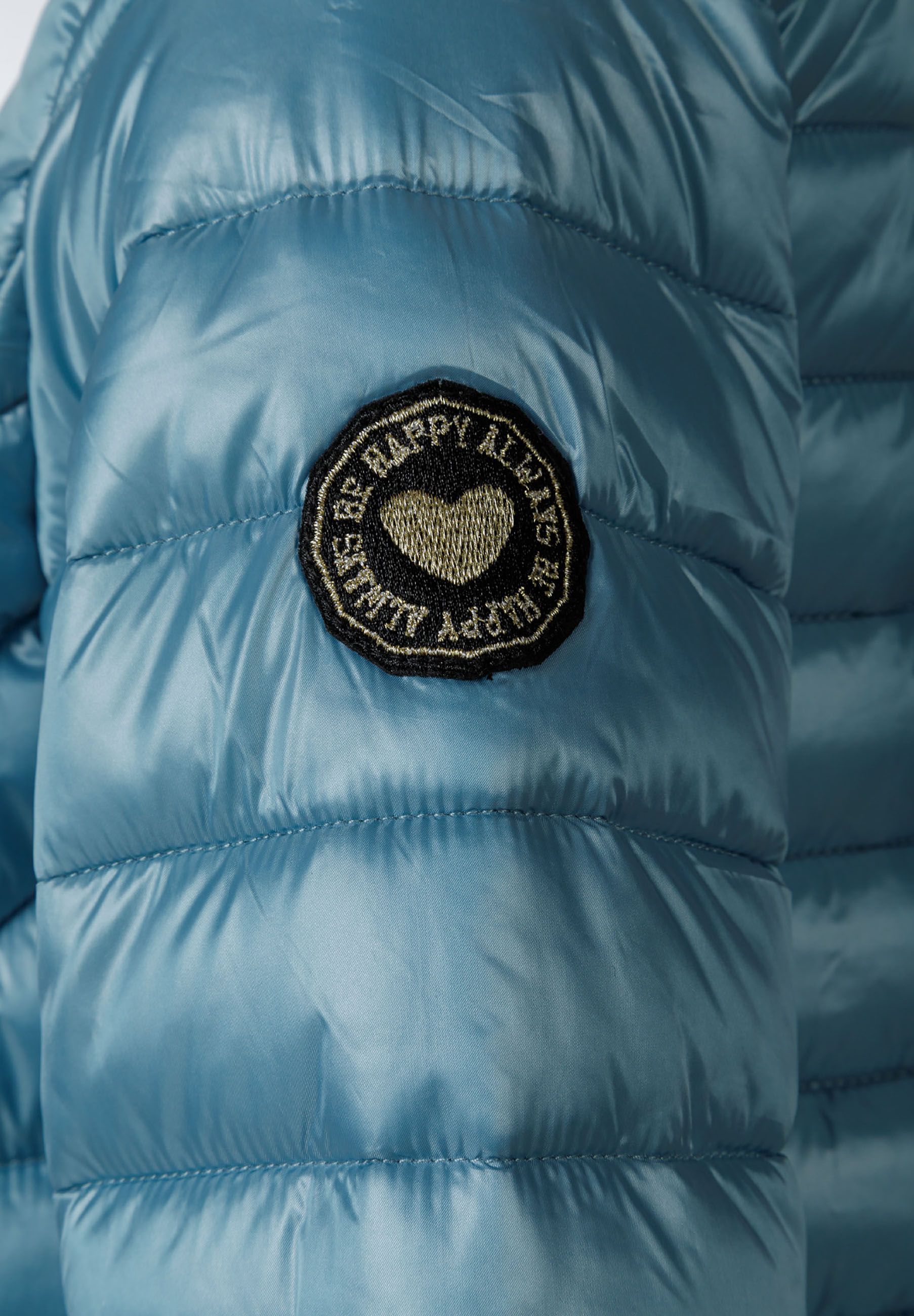 Detail of a girls quilted hooded jacket in turquoise with a heart-shaped badge.