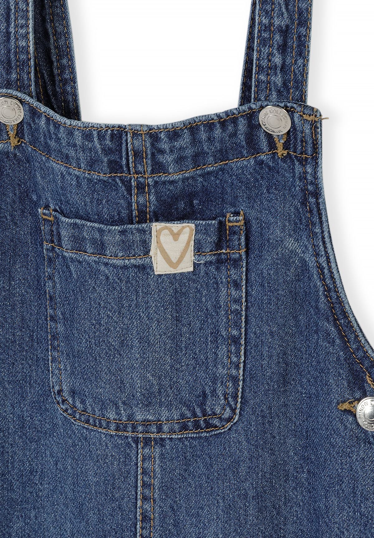 Close-up of denim dungarees bib pocket with a pale heart tab, metal strap buttons and contrast stitching.