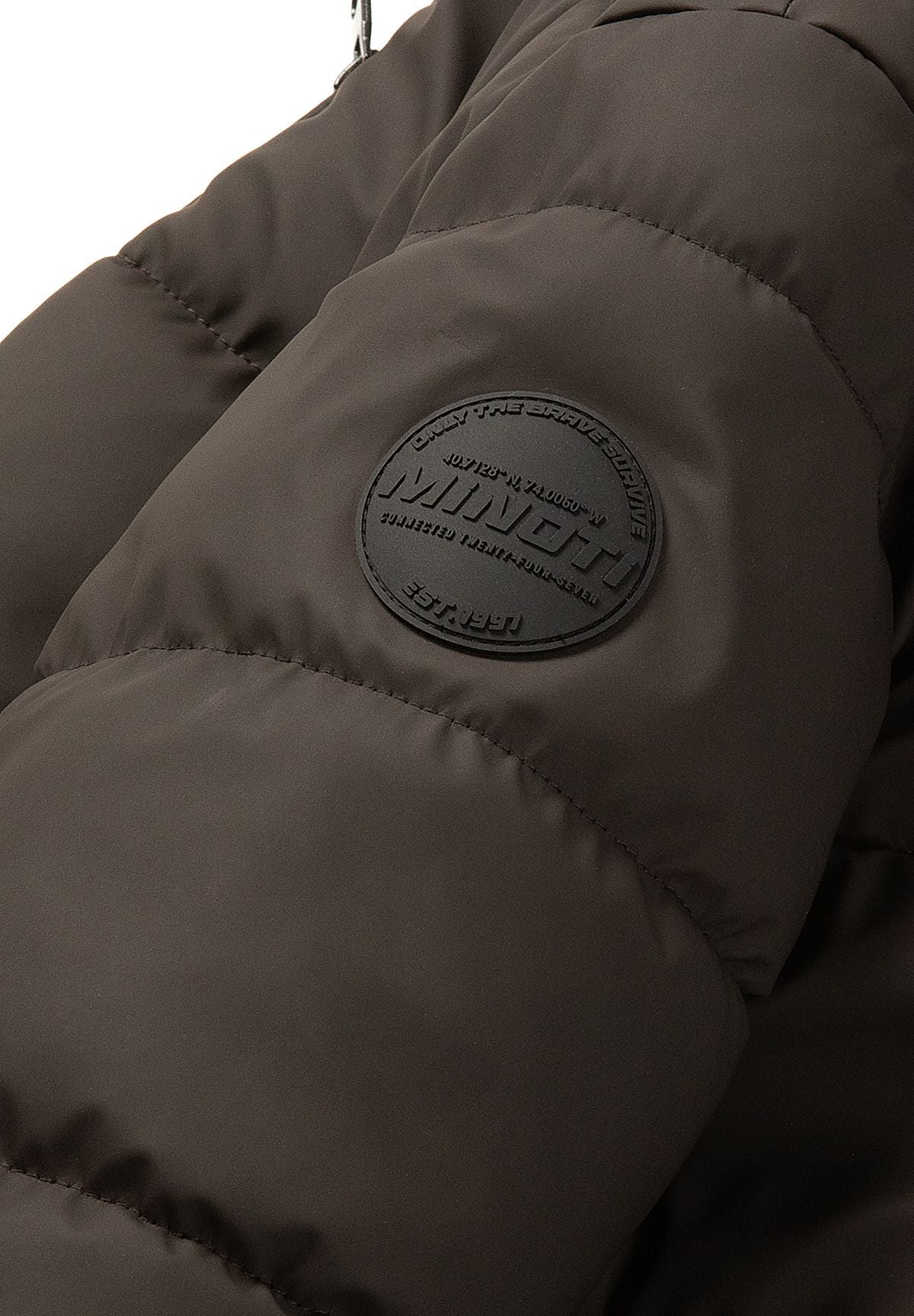 Close-up of a round dark MINOTI badge on the sleeve of a brown quilted jacket.