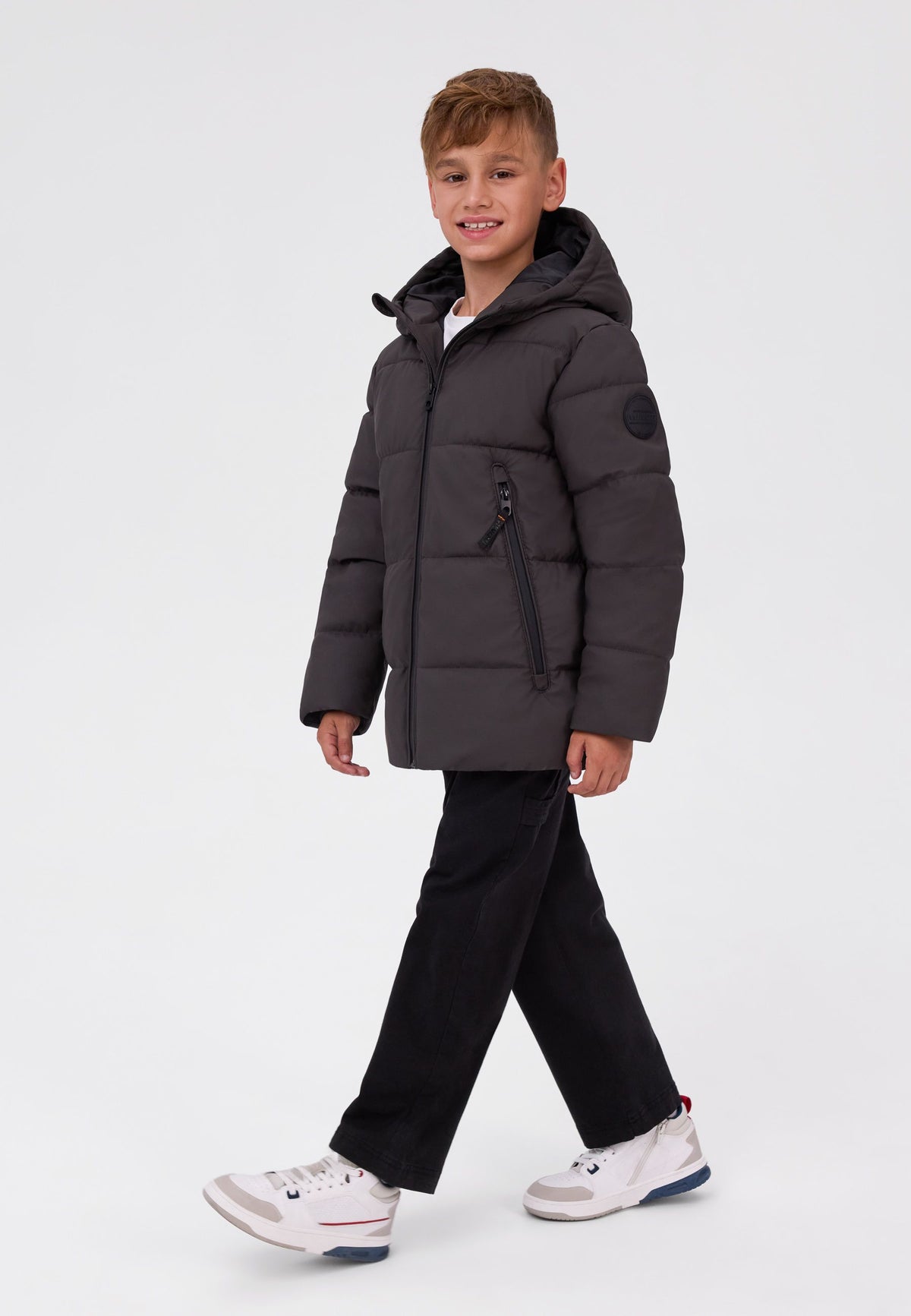 Boy wearing a dark brown hooded puffer jacket with long front zip pockets and a sleeve badge.