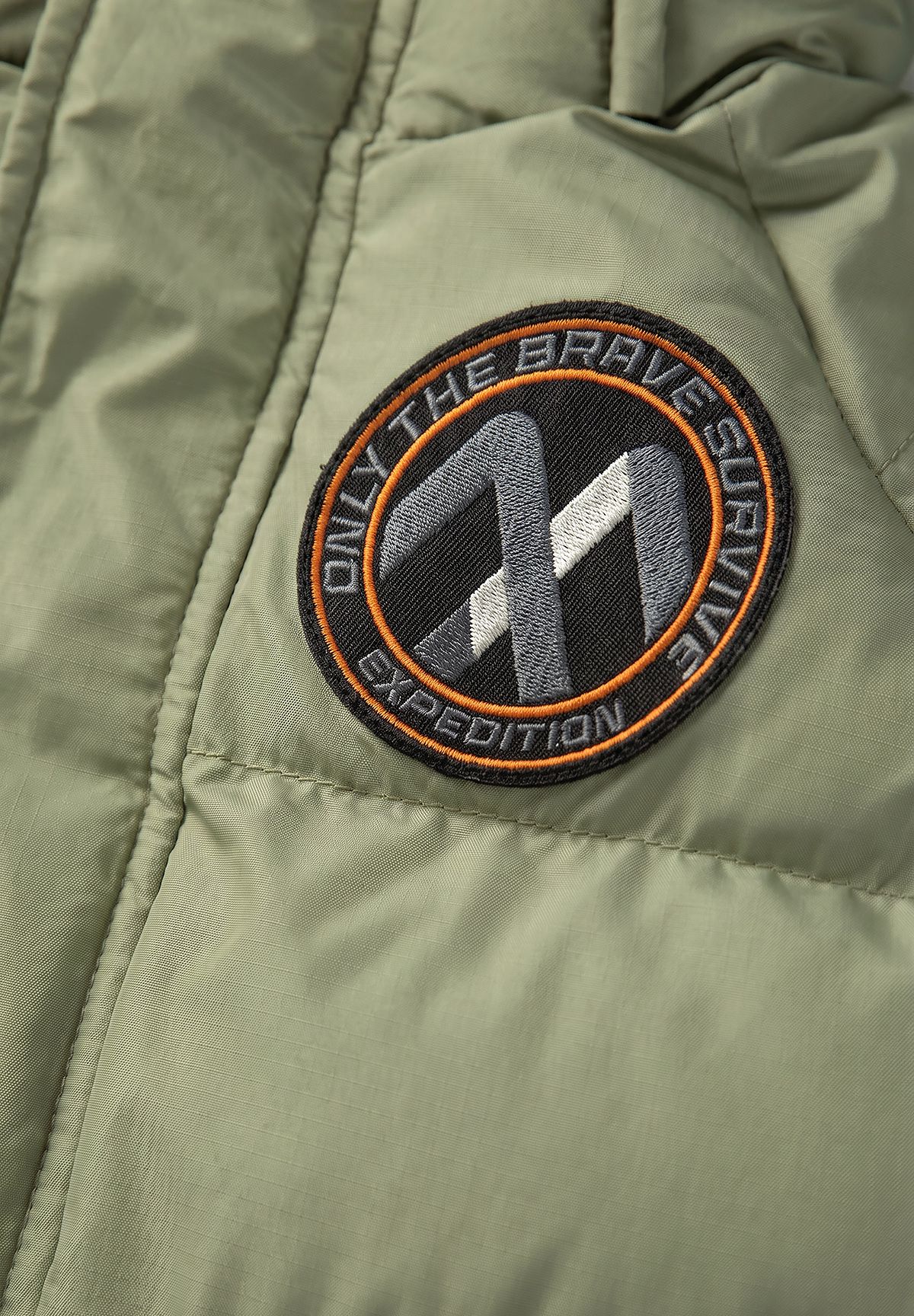 Parka acolchada de color oliva para niño con ribete de la capucha desmontable; primer plano de la insignia de la manga.