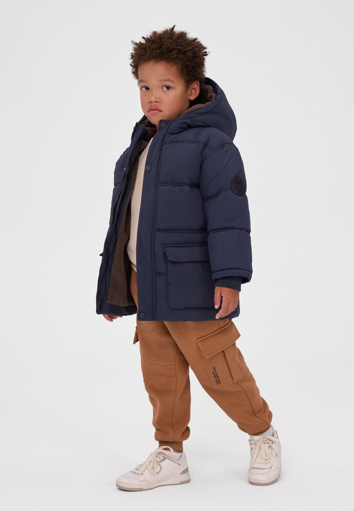 Boy wearing a navy padded parka with sherpa-lined hood, flap pockets and round sleeve patch.