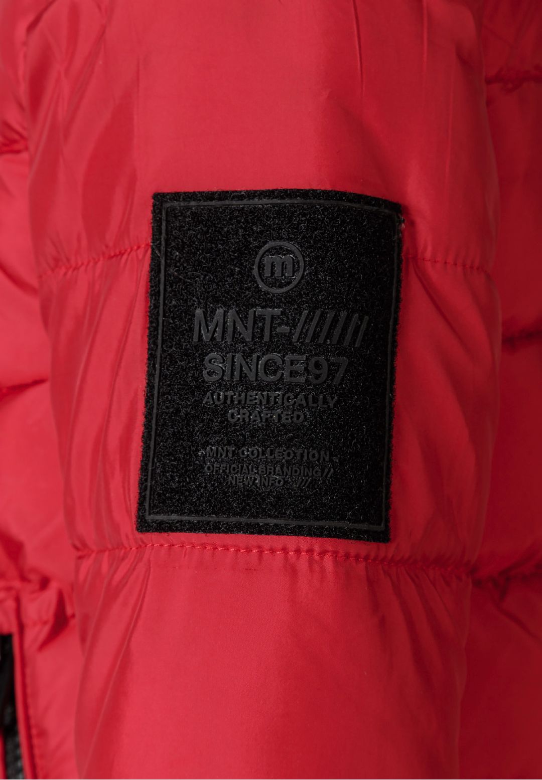 Close-up of a black rectangular MNT badge on the sleeve of the red padded jacket.