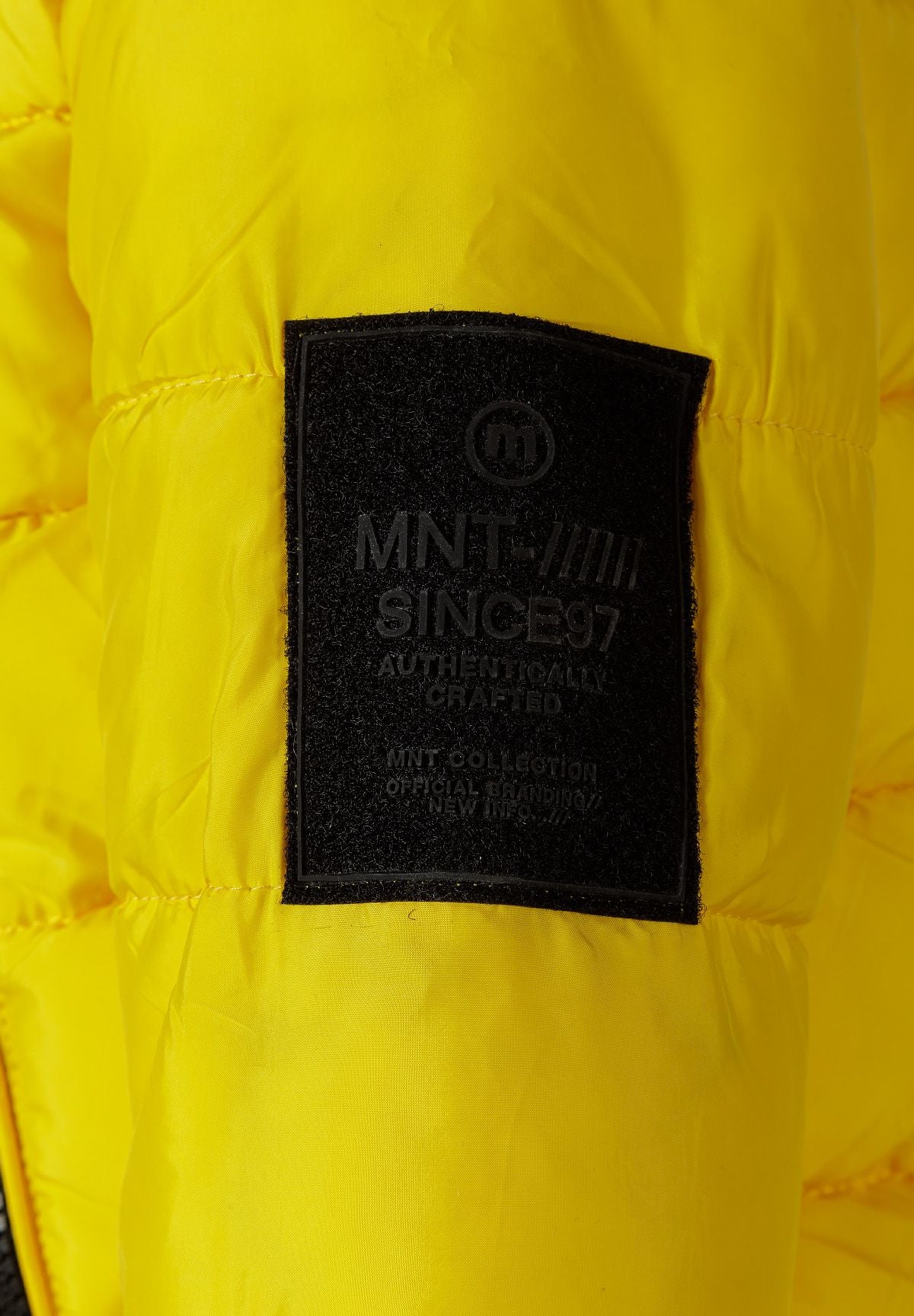 Close-up of a black MNT Since 97 rectangular patch on the yellow jacket sleeve.