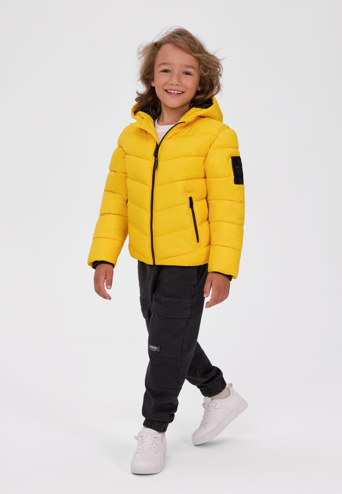 Boy wearing a bright yellow hooded puffer jacket with black zip pockets and sleeve badge.