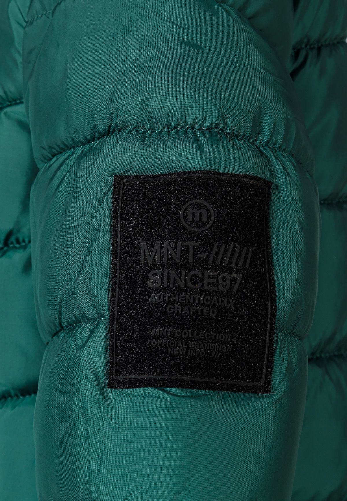 Dark rectangular MNT Since 97 badge on the teal-green padded jacket sleeve.