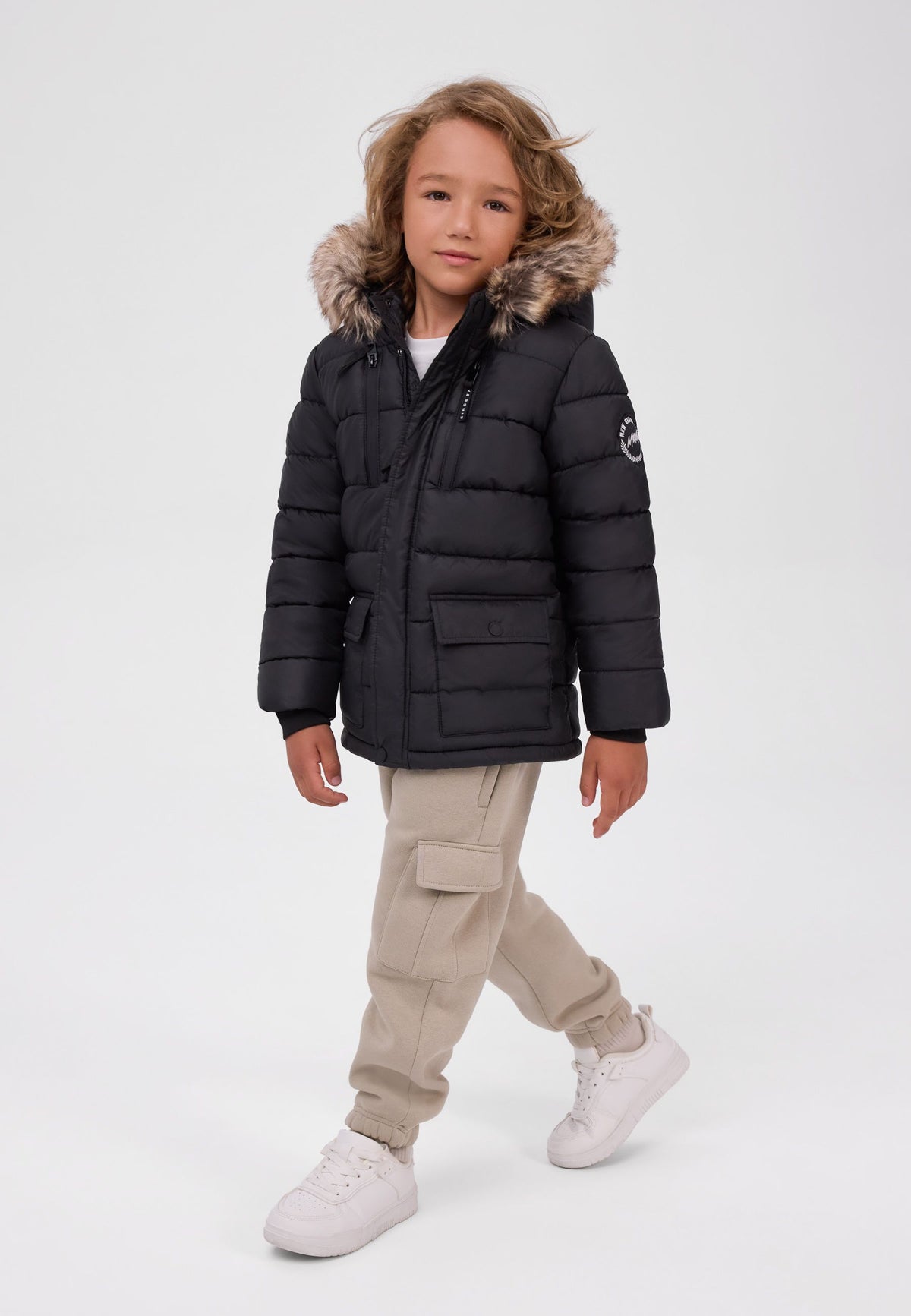 Boy wearing a black quilted parka with brown faux-fur-trimmed hood and front flap pockets.