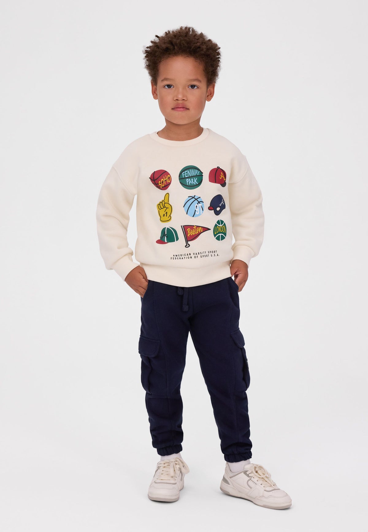 Child wearing the cream sports-print sweatshirt with navy cargo joggers and white trainers.