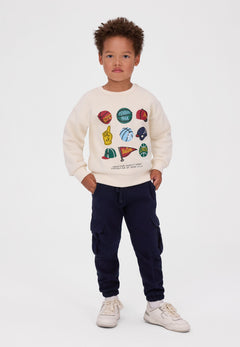 Child wearing the cream sports-print sweatshirt with navy cargo joggers and white trainers.