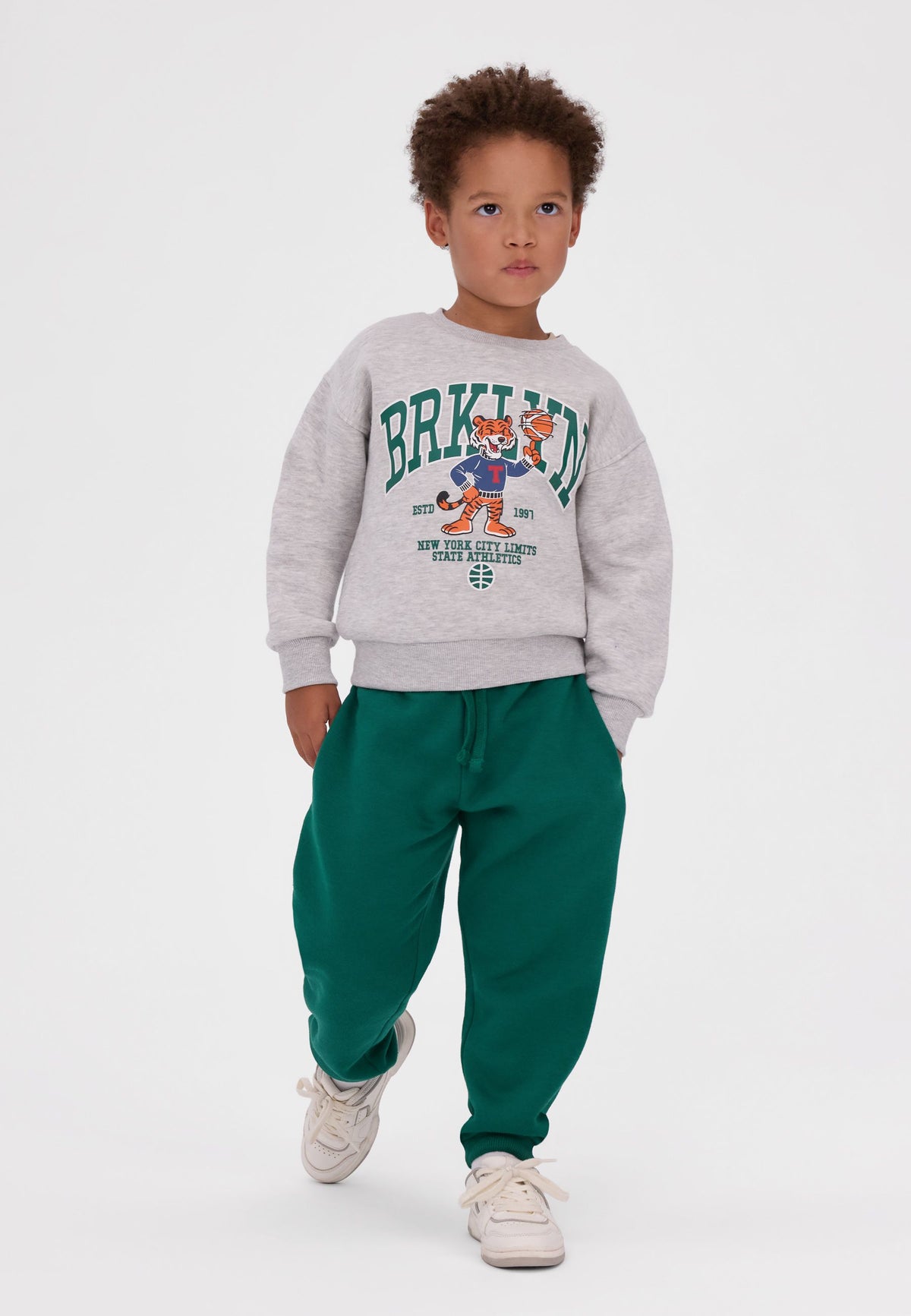 Child wearing a light grey Brooklyn tiger sweatshirt with green joggers and white trainers.