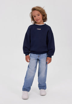 Child wearing a navy Studio Dept sweatshirt with light blue jeans and white trainers.