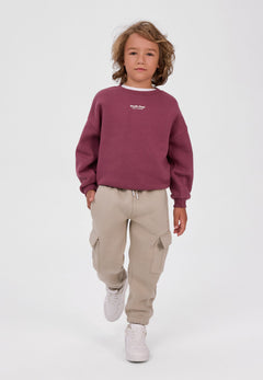 Child wearing burgundy Studio Dept sweatshirt with beige cargo joggers and white trainers.