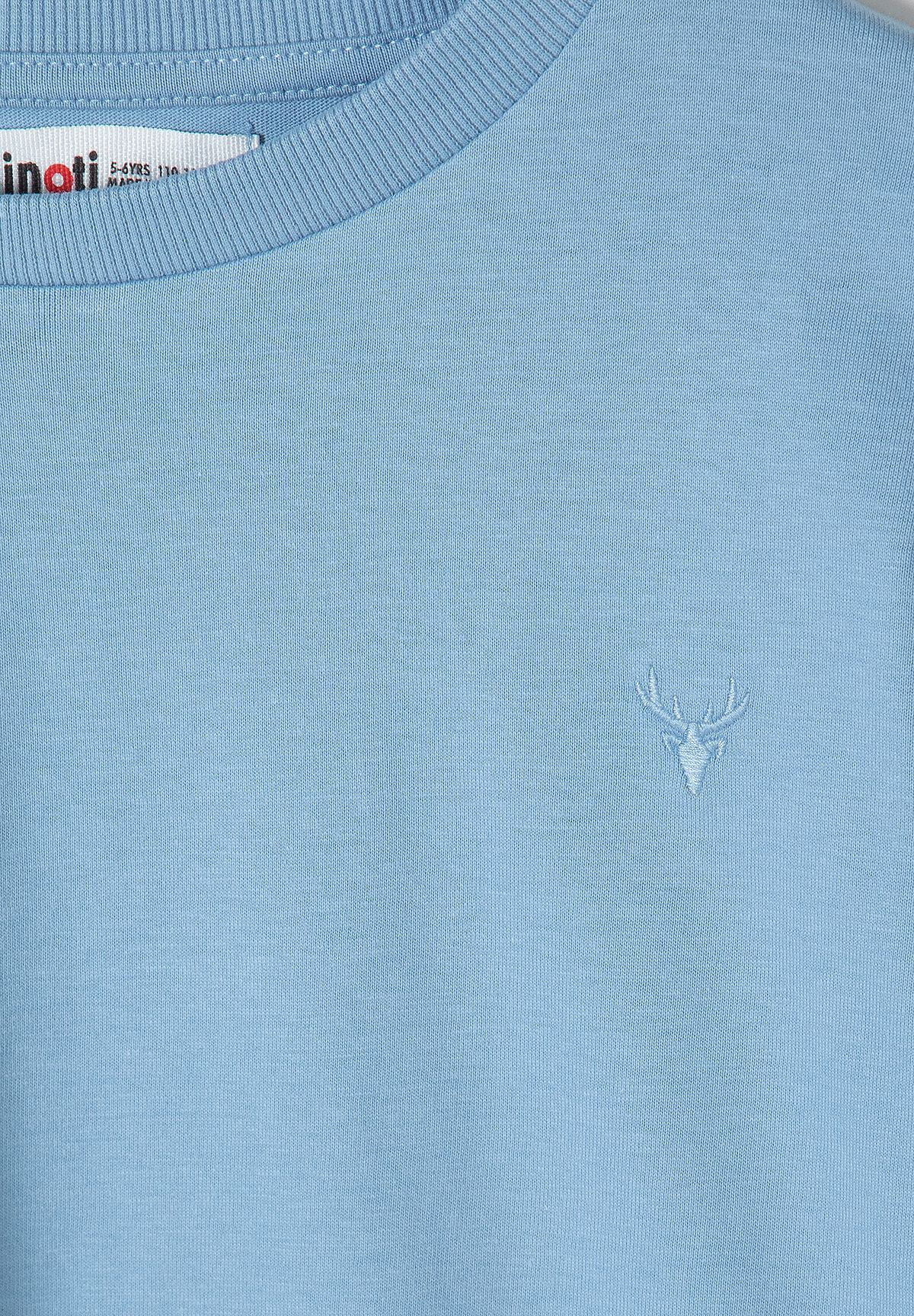 Close-up of small white stag-head embroidery on light blue top beneath ribbed crew neck.