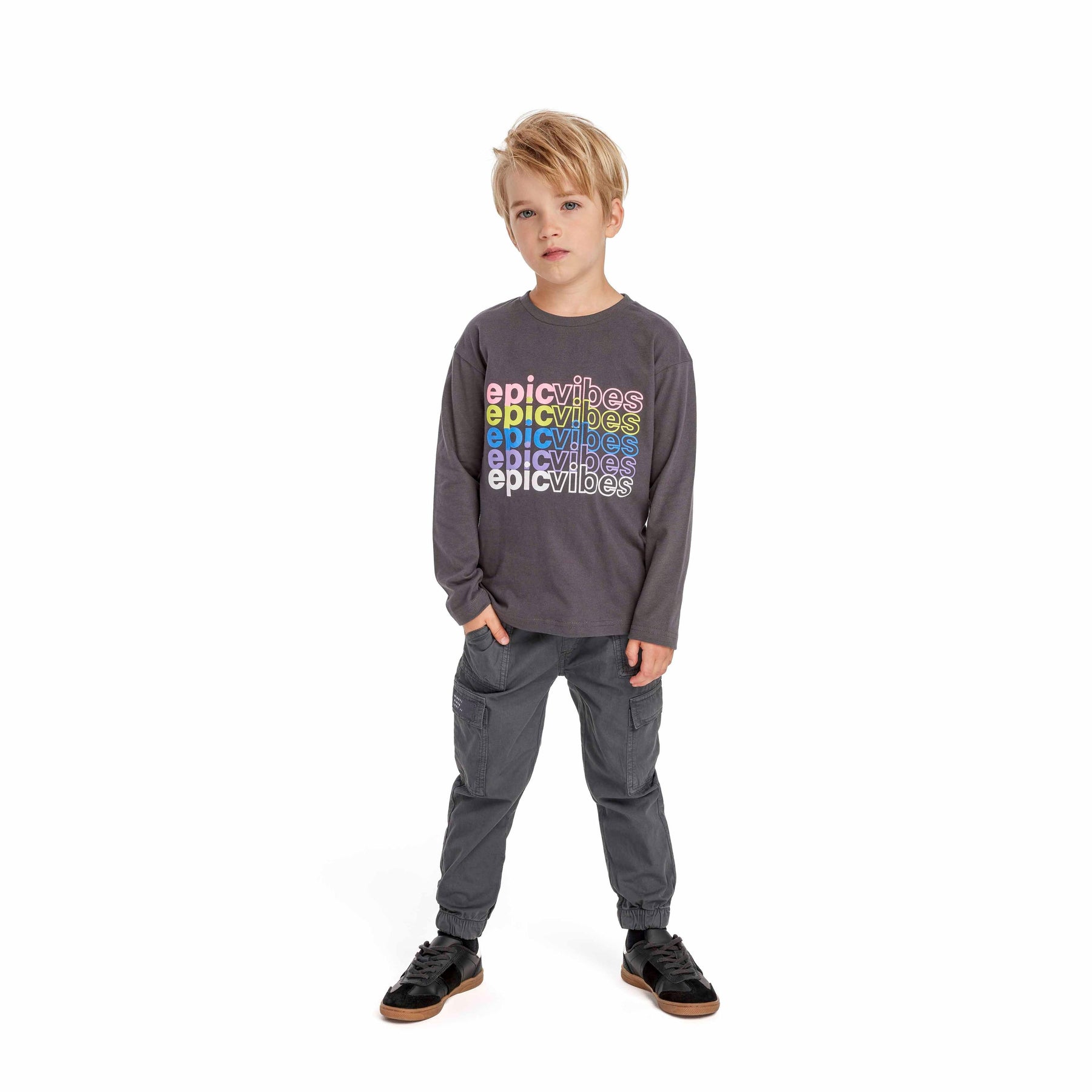 Young blonde boy wearing black combat trousers and grey long-sleeve top with colorful text, standing against white background with hands in pockets